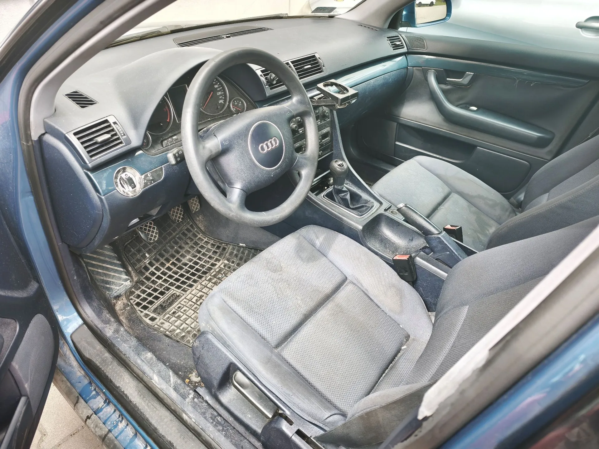 Wnętrze samochodu marki Audi widziane z perspektywy otwartych drzwi kierowcy. Widoczna jest ciemnoszara kierownica z logo czterech pierścieni, deska rozdzielcza z zegarami, lewarek zmiany biegów oraz dwa przednie fotele z materiałowej tapicerki w kolorze szarym. Na podłodze znajduje się gumowa mata z wypustkami.