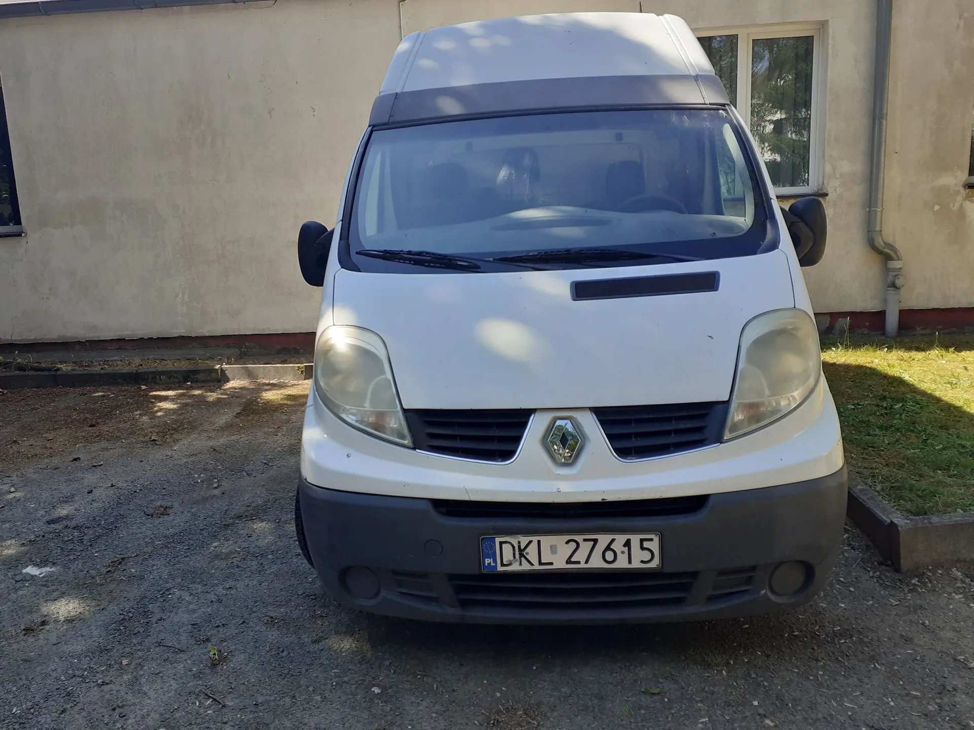 Biały samochód dostawczy marki Renault zaparkowany przodem do obiektywu na żwirowym podjeździe przed jasnym budynkiem. Pojazd posiada wysoką nadbudowę dachu oraz tablicę rejestracyjną o numerze DKL 27615.
