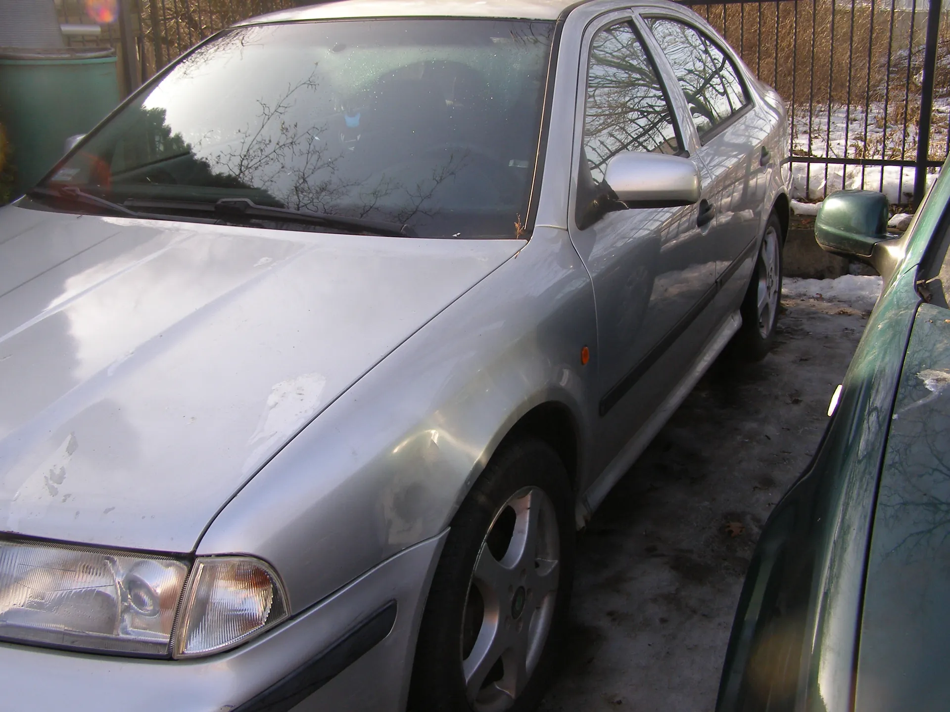 Srebrny samochód osobowy typu sedan zaparkowany na zimowym podwórku. Widoczna przód i bok pojazdu z reflektorami, lusterkiem bocznym oraz oponami. Obok, po prawej stronie, fragment zielonego samochodu. Tło: metalowe ogrodzenie, suche drzewa bez liści, śnieg na ziemi oraz zielony pojemnik. Podłoże częściowo pokryte śniegiem i lodem.