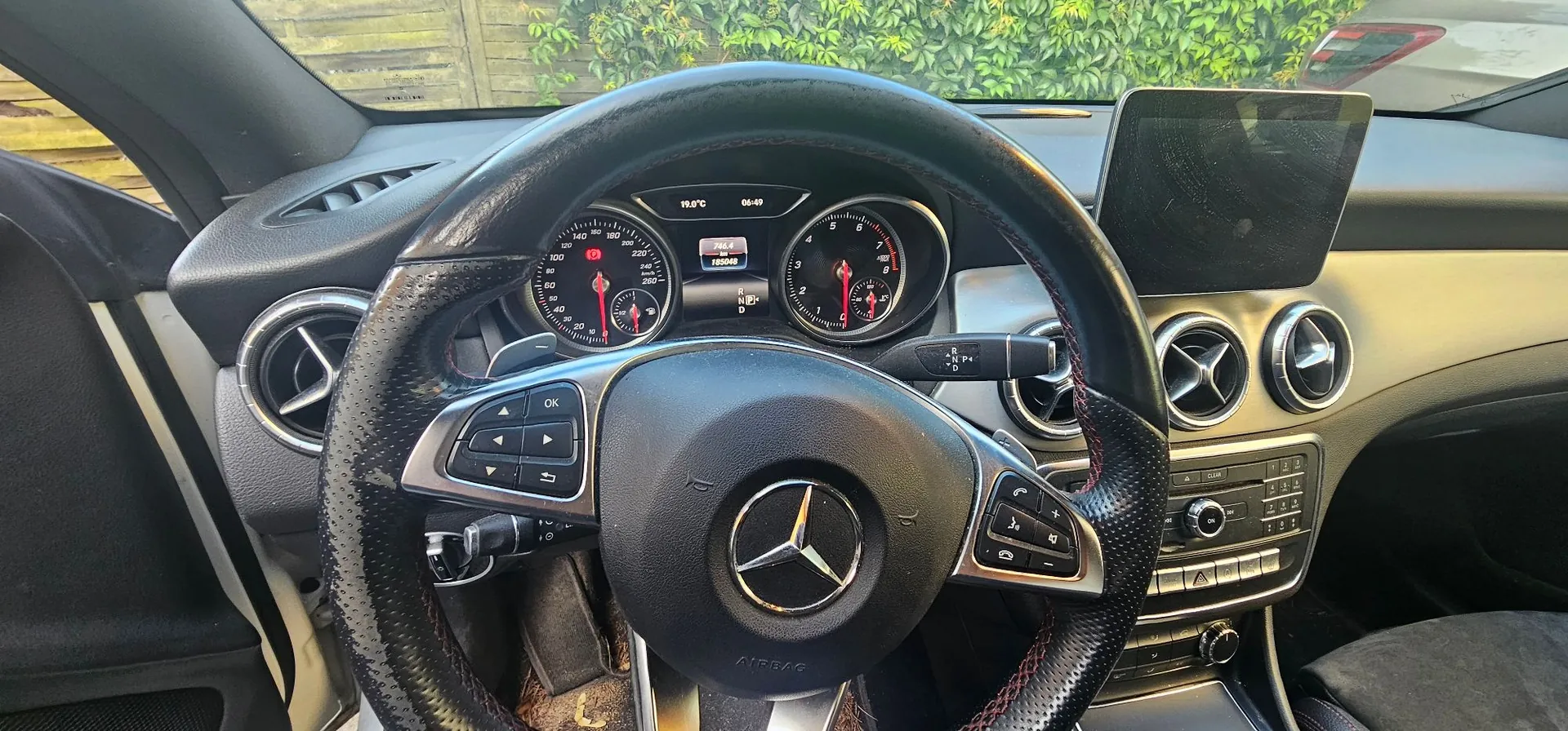 Widok z wnętrza samochodu marki Mercedes-Benz z perspektywy kierowcy. W centrum znajduje się wielofunkcyjna kierownica obszyta czarną skórą z karbonowymi wstawkami i logo Mercedesa na środku. Za kierownicą widoczny jest zestaw wskaźników z dwoma dużymi analogowymi zegarami (prędkościomierzem i obrotomierzem) oraz małym wyświetlaczem cyfrowym w centrum, na którym widnieje temperatura 19.5°C i godzina 06:49. Po prawej stronie znajduje się ekran multimedialny oraz panel centralny z przyciskami i okrągłymi nawiewami powietrza.