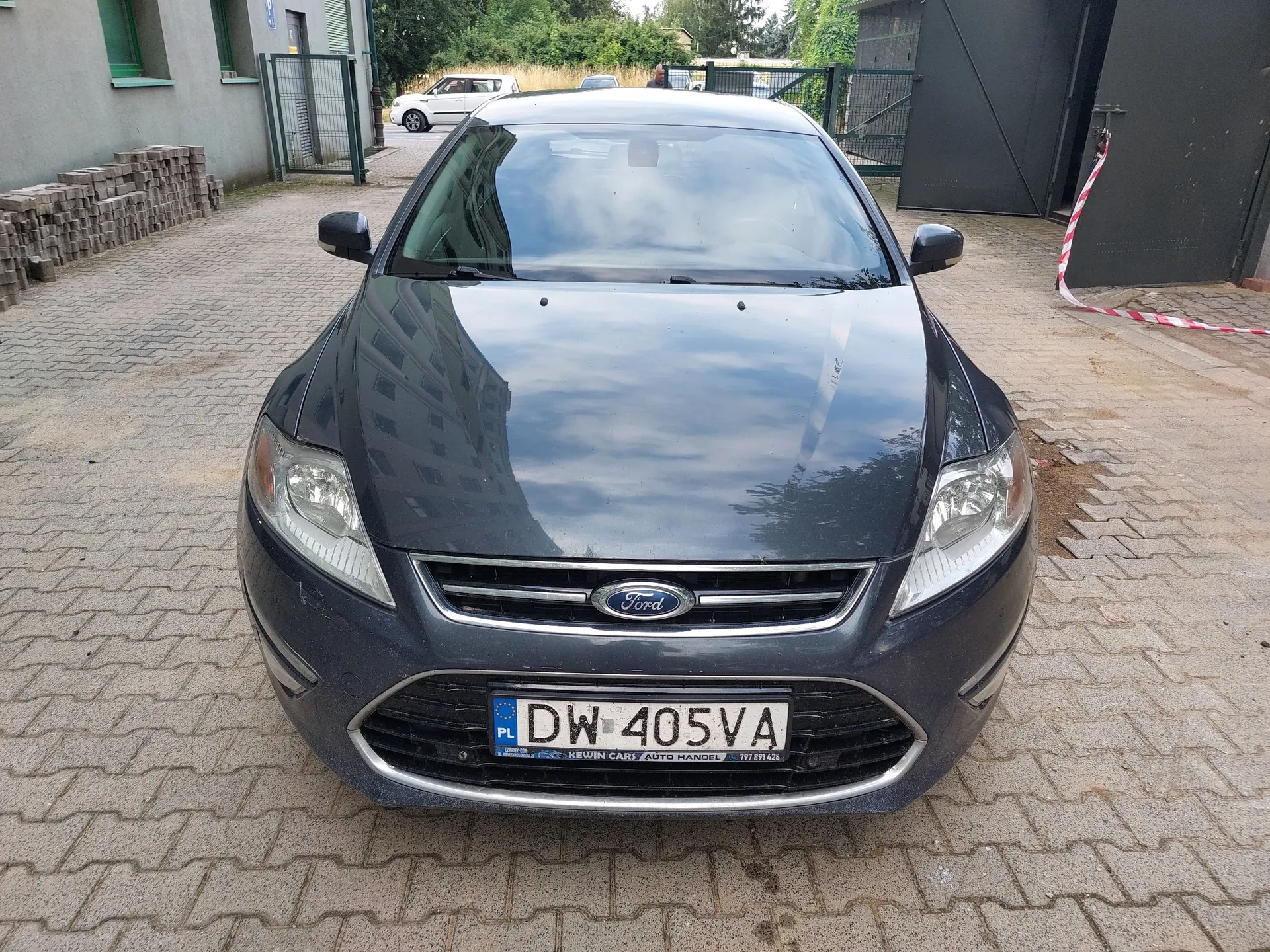 Widok z przodu na ciemnoszary samochód marki Ford zaparkowany na kostce brukowej. Pojazd posiada polską tablicę rejestracyjną o numerze DW 405VA. Poniżej numeru rejestracyjnego znajduje się napis „KEWIN CARS AUTO HANDEL 777 611 426”. W tle widoczny jest fragment budynku, ogrodzenie oraz biały samochód zaparkowany w oddali.