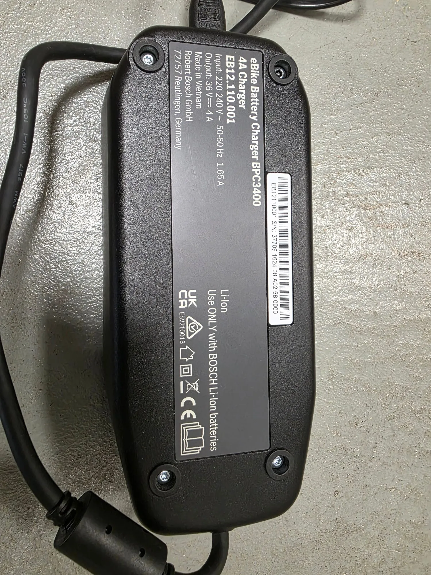 Czarna ładowarka do baterii ebike Bosch BPC3400. Na obudowie znajduje się biały tekst: 'eBike Battery Charger BPC3400', 'EB12110001', 'Input: 220-240V ~ 50-60Hz 1.65A', 'Output: 36V ⎓ 4A', 'Made in Vietnam', 'Robert Bosch GmbH 72757 Reutlingen, Germany'. Widoczny jest kod kreskowy z numerem seryjnym S/N: 37709 1824 08 A02 59 0000 oraz oznaczenia: 'Li-Ion', 'Use ONLY with BOSCH Li-Ion batteries', znaki certyfikacji UKCA, CE oraz symbole utylizacji. Urządzenie ma dwa czarne kable wychodzące z przeciwległych końców.
