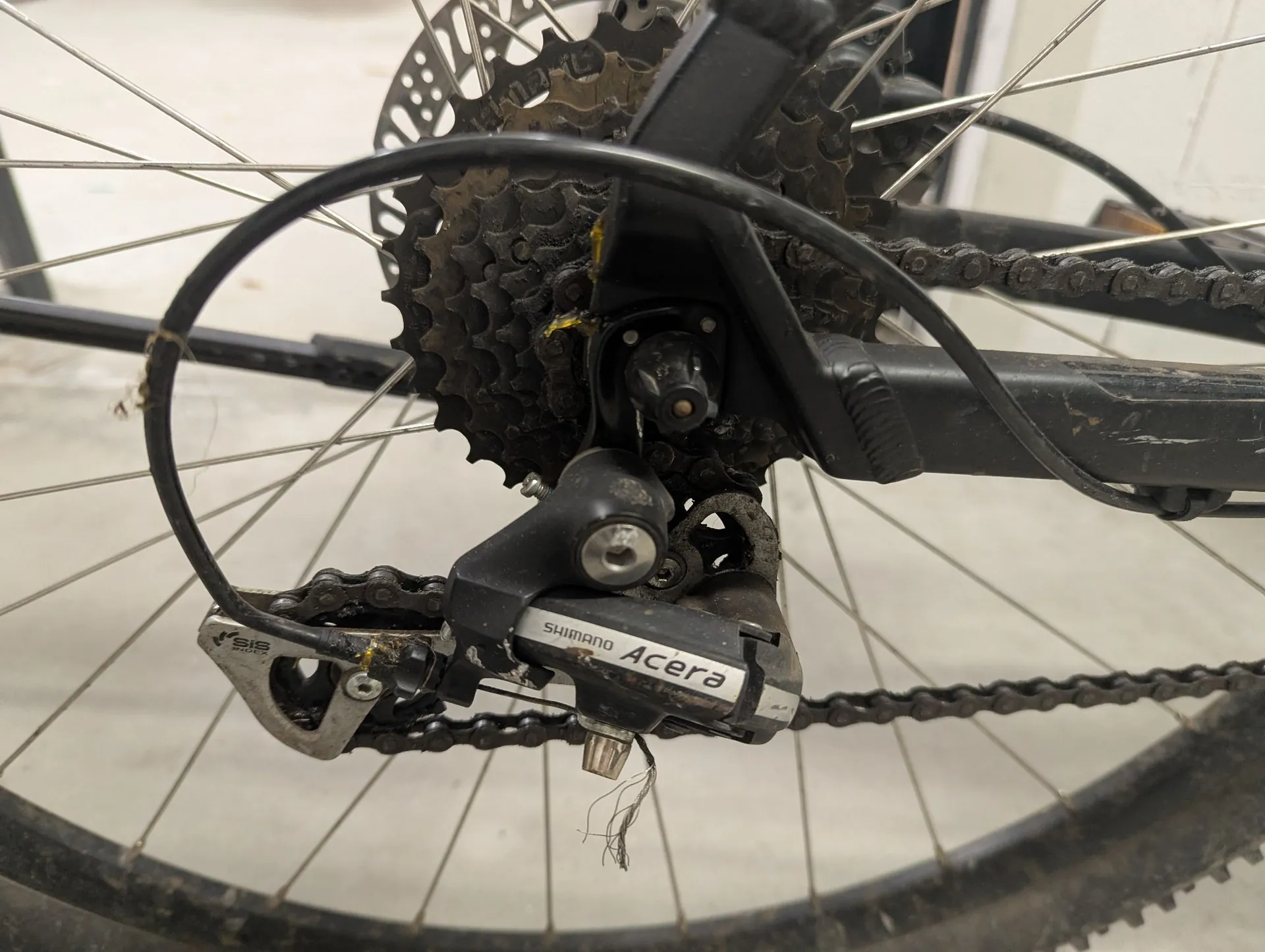 Zbliżenie na tylną przerzutkę i kasetę roweru. Widoczny jest napęd z łańcuchem, czarne zębatki kasety oraz przerzutka marki Shimano z napisem 'SHIMANO Acera' oraz 'SIS'. Elementy są lekko zabrudzone smarem i kurzem. W tle widoczne są szprychy koła i fragment ramy roweru.