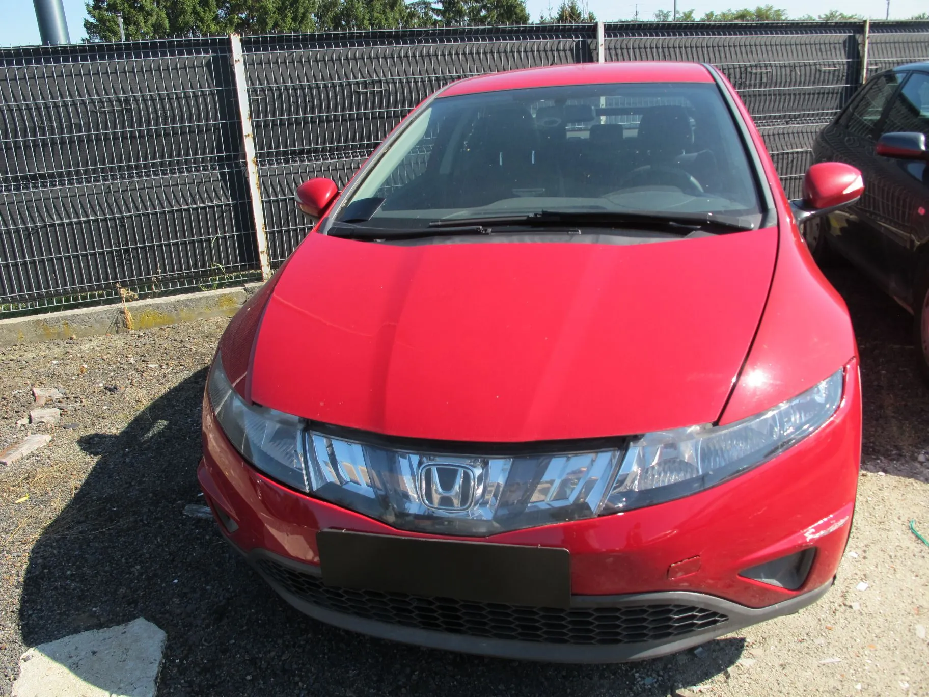 Czerwony samochód marki Honda widziany od przodu, zaparkowany na żwirowym placu przed czarnym ogrodzeniem z siatki. Widoczny jest emblemat Hondy na przednim grillu oraz czarna tablica rejestracyjna bez tekstu.
