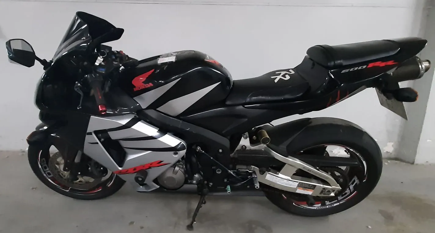 Czarny motocykl sportowy marki Honda z srebrnymi i czerwonymi akcentami, zaparkowany bokiem na jasnym tle w pomieszczeniu. Na baku widoczne jest logo Hondy, a na tylnej części siedzenia napis 'CBR'. Motocykl stoi na bocznej stopce.