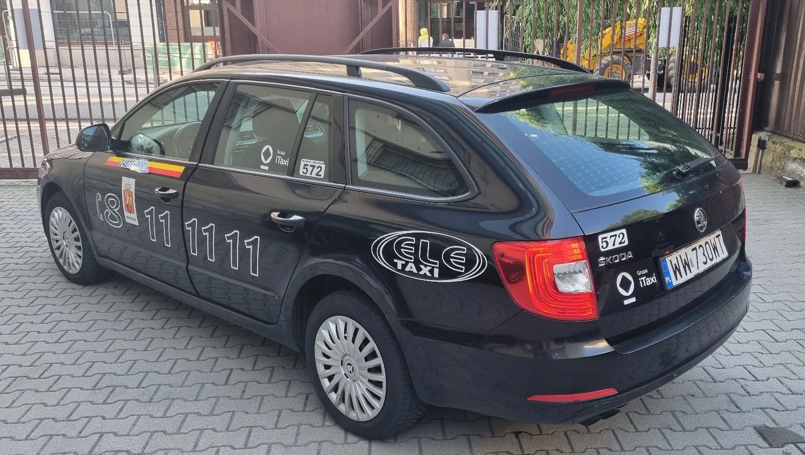 Czarny samochód marki Skoda w wersji kombi, pełniący funkcję taksówki, zaparkowany na kostce brukowej. Na boku pojazdu widnieje duży biały numer telefonu '8 11 11 11' oraz logo 'ELE TAXI'. Na tylnej klapie znajduje się tablica rejestracyjna 'WW 730WT', logo 'ŠKODA' oraz napis 'Grupa iTaxi'. Na bocznej szybie widnieje naklejka z numerem '572'.