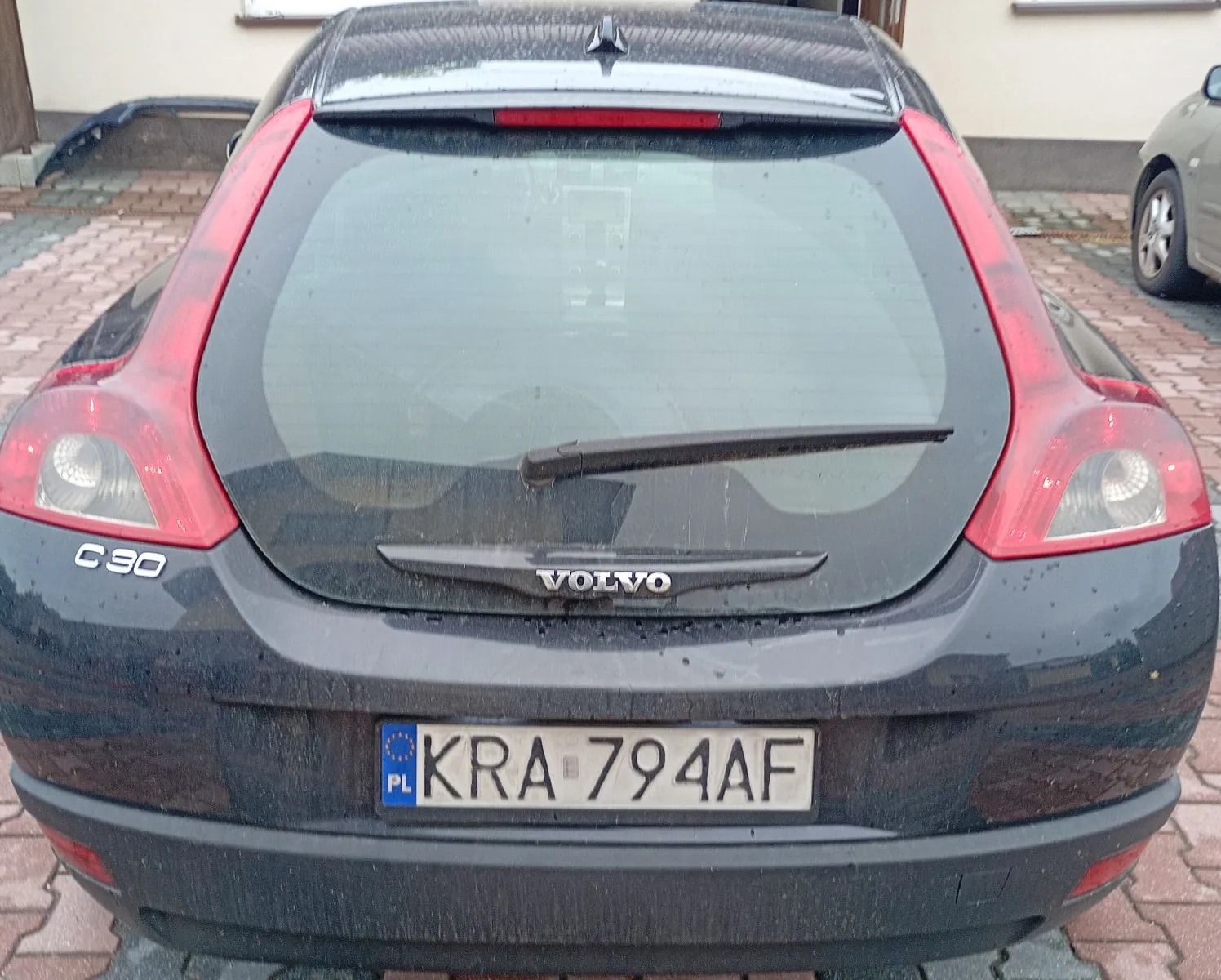 Tylny widok ciemnoszarego samochodu marki Volvo, model C30. Na klapie bagażnika widnieje napis VOLVO oraz oznaczenie modelu C30 po lewej stronie. Samochód ma polską tablicę rejestracyjną o numerze KRA 794AF. Karoseria jest lekko zabrudzona.
