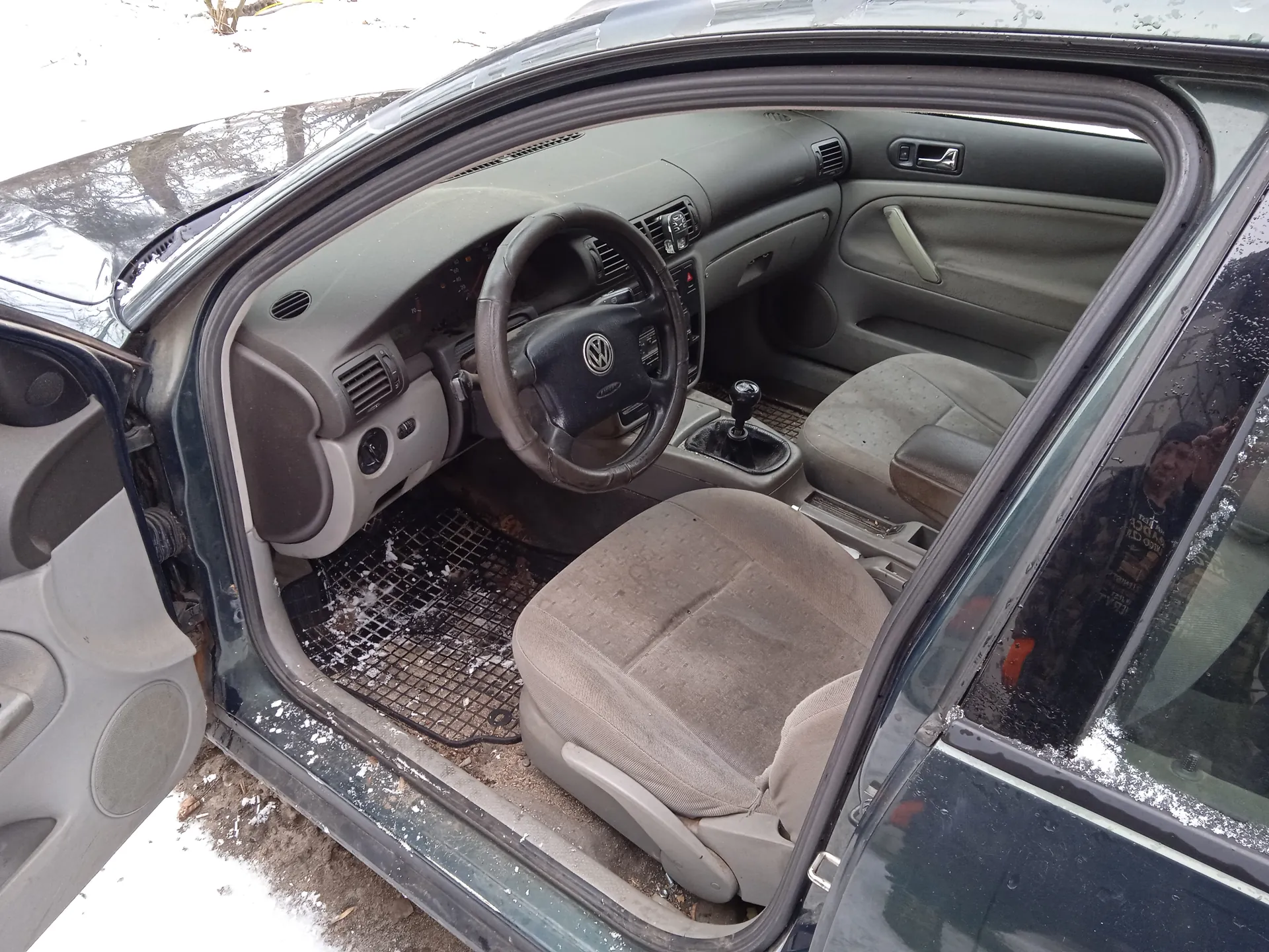 Wnętrze samochodu marki Volkswagen z otwartą przodem drzwiami od strony kierowcy. Widoczna kierownica z logo VW, szare siedzenia, deska rozdzielcza, dźwignia zmiany biegów oraz dywaniki z siatką na podłodze. Na dywanikach i podłodze wokół samochodu znajduje się śnieg. Zewnętrzna część pojazdu jest ciemnoszara. W tle widoczne są drzewa i śnieżna powierzchnia. W lusterku bocznym odbija się postać człowieka.