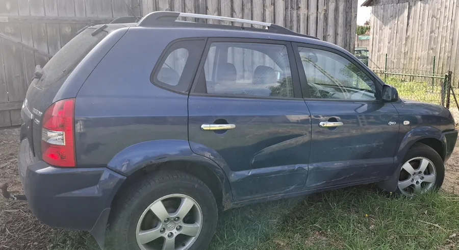 Ciemnoniebieski SUV zaparkowany na trawie obok drewnianego ogrodzenia. Samochód posiada dachowy bagażnik, srebrne uchwyty drzwiowe i felgi aluminiowe. Widoczne są uszkodzenia na boku nadwozia. W tle drewniana płot i brama.