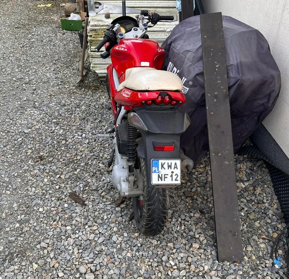 Czerwony motocykl marki Gilera zaparkowany na żwirowym podłożu. Widoczna tablica rejestracyjna z napisem PL KWA NF12. Na siedzeniu znajduje się beżowa pokrywa. Po prawej stronie motocykla stoi przedmiot zakryty ciemnym pokrowcem z częściowo widocznym napisem. W tle stos drewnianych desek, zielone pudełko oraz metalowa belka oparta o ścianę. Podłoże pokryte jest drobnym żwirem.