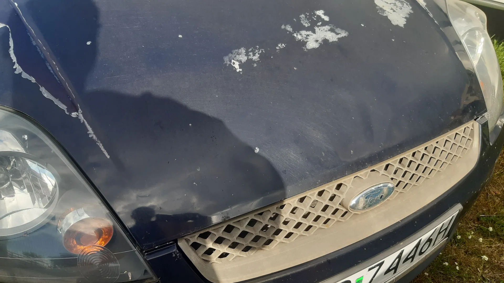 Zbliżenie na przód ciemnoniebieskiego samochodu marki Ford. Na masce widoczne są liczne uszkodzenia lakieru w postaci białych odprysków i zmatowień. Widoczny jest fragment reflektora po lewej stronie, grill z logo Ford oraz fragment tablicy rejestracyjnej z widocznymi cyframi i literami 7446H.