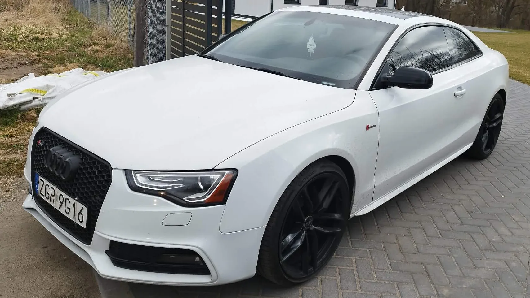 Biały samochód marki Audi w nadwoziu coupe, zaparkowany na kostce brukowej. Pojazd ma czarne felgi, czarną atrapę chłodnicy w kształcie plastra miodu oraz tablicę rejestracyjną o numerze ZGR 9G16. W tle widoczny jest fragment ogrodzenia i trawnik.