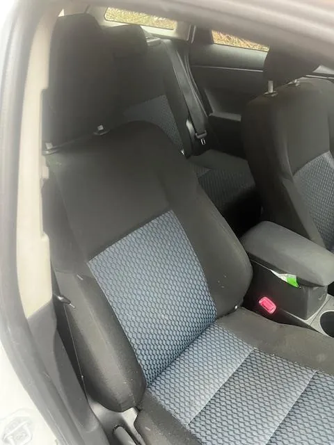 Wnętrze samochodu z widocznymi czarnymi fotelami z niebieskim wzorzystym materiałem. Widoczne są przednie i tylne siedzenia, otwarte drzwi oraz fragment zewnętrzny z trawą za oknem. W centralnej konsoli znajduje się różowy element i zielony przedmiot. Siedzenia mają czarne poduszki z niebieskimi wstawkami w geometryczny wzór.