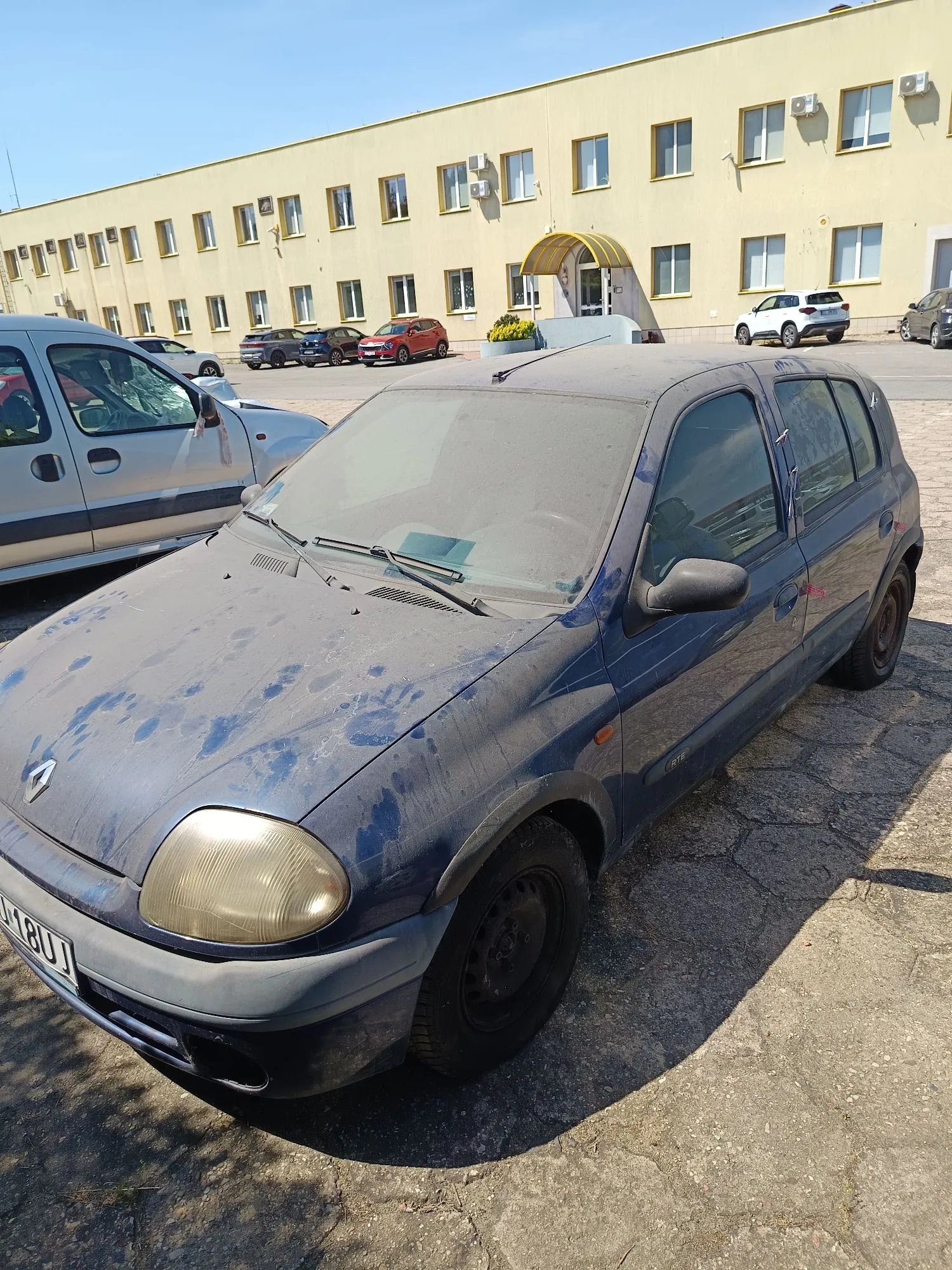Niebieski, zakurzony samochód osobowy marki Renault zaparkowany na betonowym placu przed żółtym, dwukondygnacyjnym budynkiem. Auto ma zmatowiałe reflektory i widoczne ślady zabrudzeń na masce. W tle widoczne są inne zaparkowane samochody oraz jasne niebo.