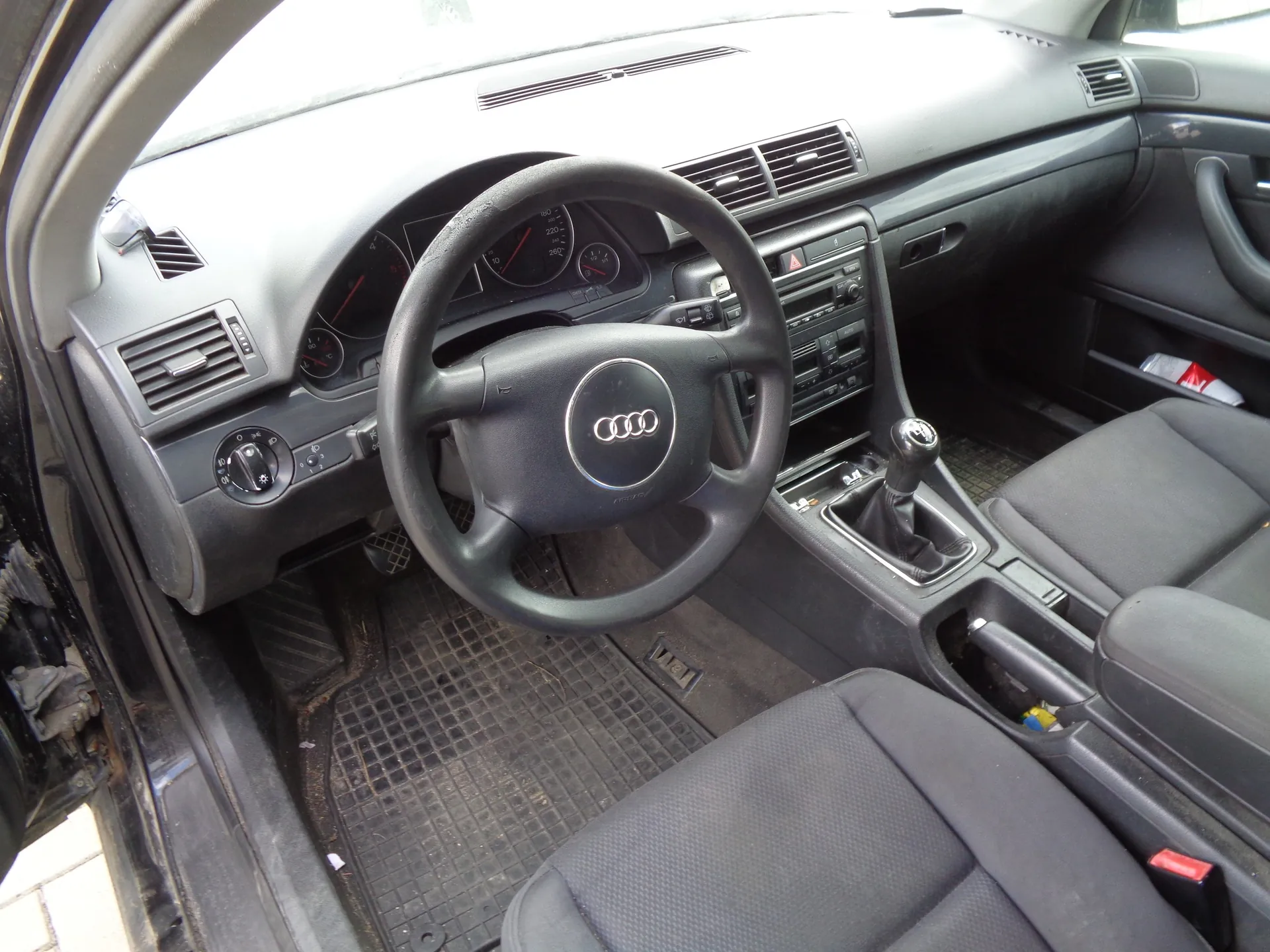 Wnętrze samochodu marki Audi, widok z perspektywy otwartych drzwi kierowcy. Widoczna czarna kierownica z logo Audi, deska rozdzielcza z zegarami, nawiewami, radiem oraz lewarem zmiany biegów. W samochodzie znajdują się ciemnoszare, materiałowe fotele oraz gumowa mata na podłodze.