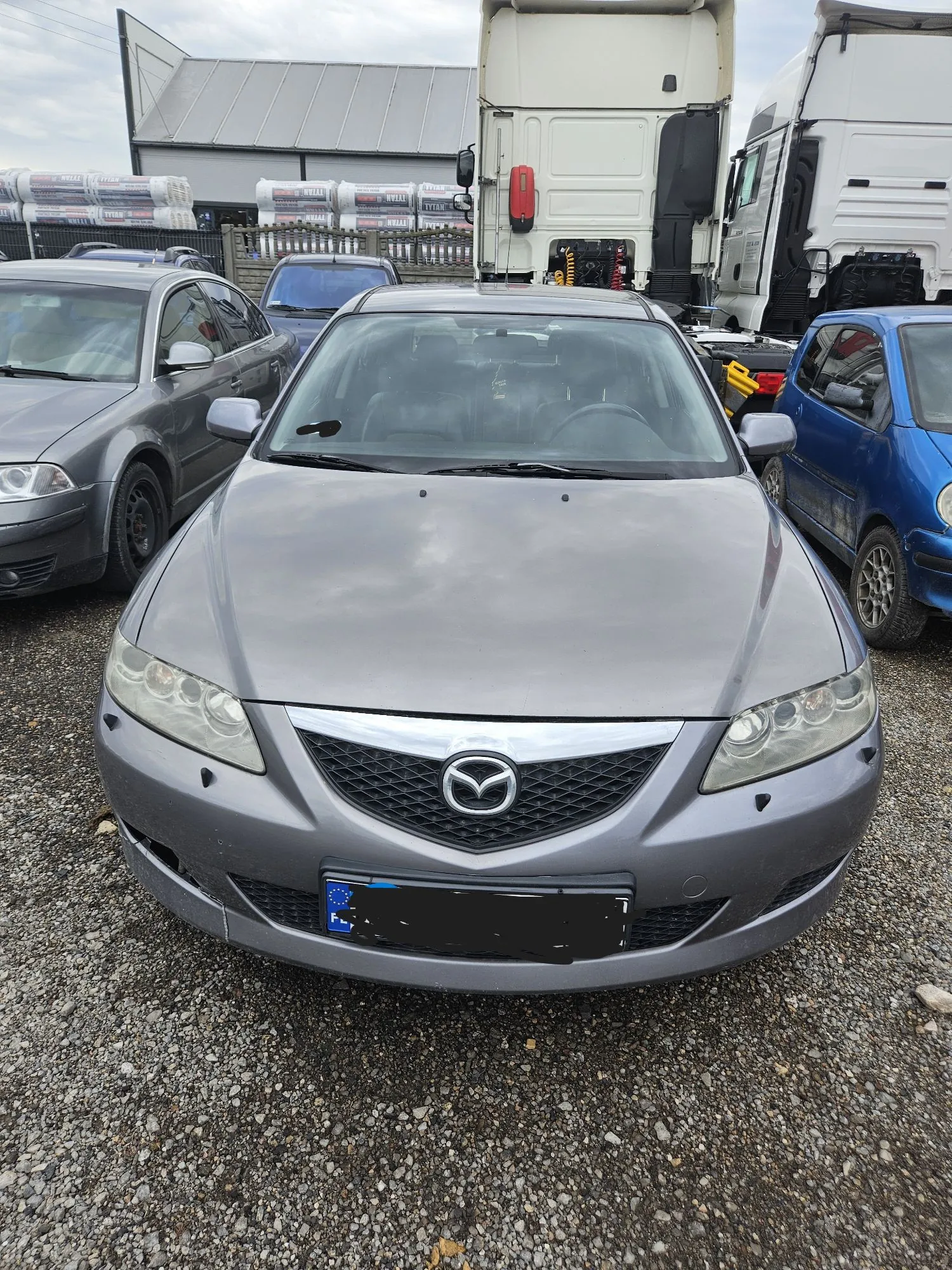 Srebrny samochód osobowy marki Mazda zaparkowany przodem do obiektywu na żwirowym placu. W tle widoczne są inne pojazdy: szary samochód po lewej stronie, niebieski samochód po prawej oraz białe ciężarówki w głębi. W oddali znajduje się budynek z szarym dachem oraz stosy materiałów budowlanych owiniętych w folię.
