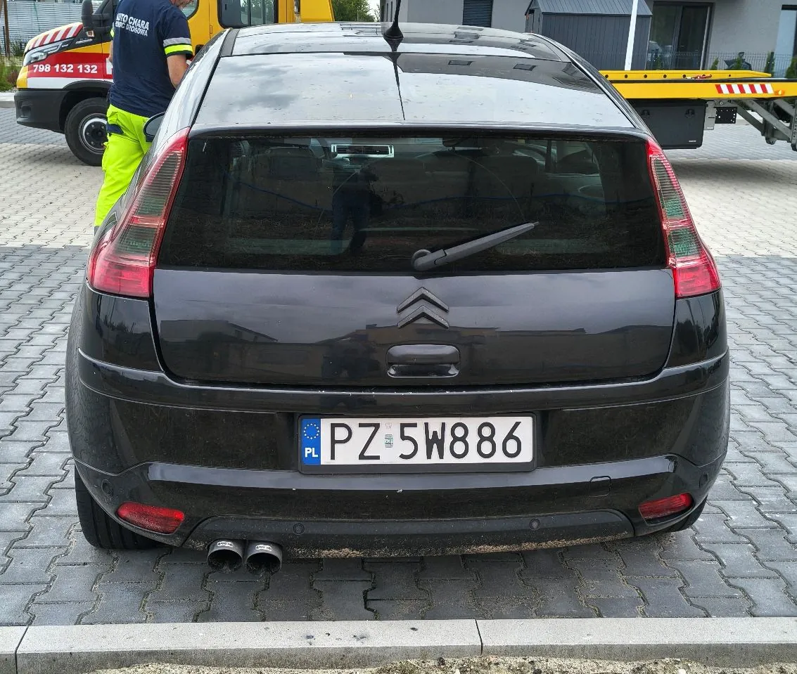 Tylny widok czarnego samochodu marki Citroën zaparkowanego na kostce brukowej. Na samochodzie widoczna jest tablica rejestracyjna o numerze PZ 5W886. W tle po lewej stronie znajduje się fragment żółto-czerwonego pojazdu assistance z numerem telefonu 798 132 132 oraz osoba w neonowej zielonej odzieży roboczej.