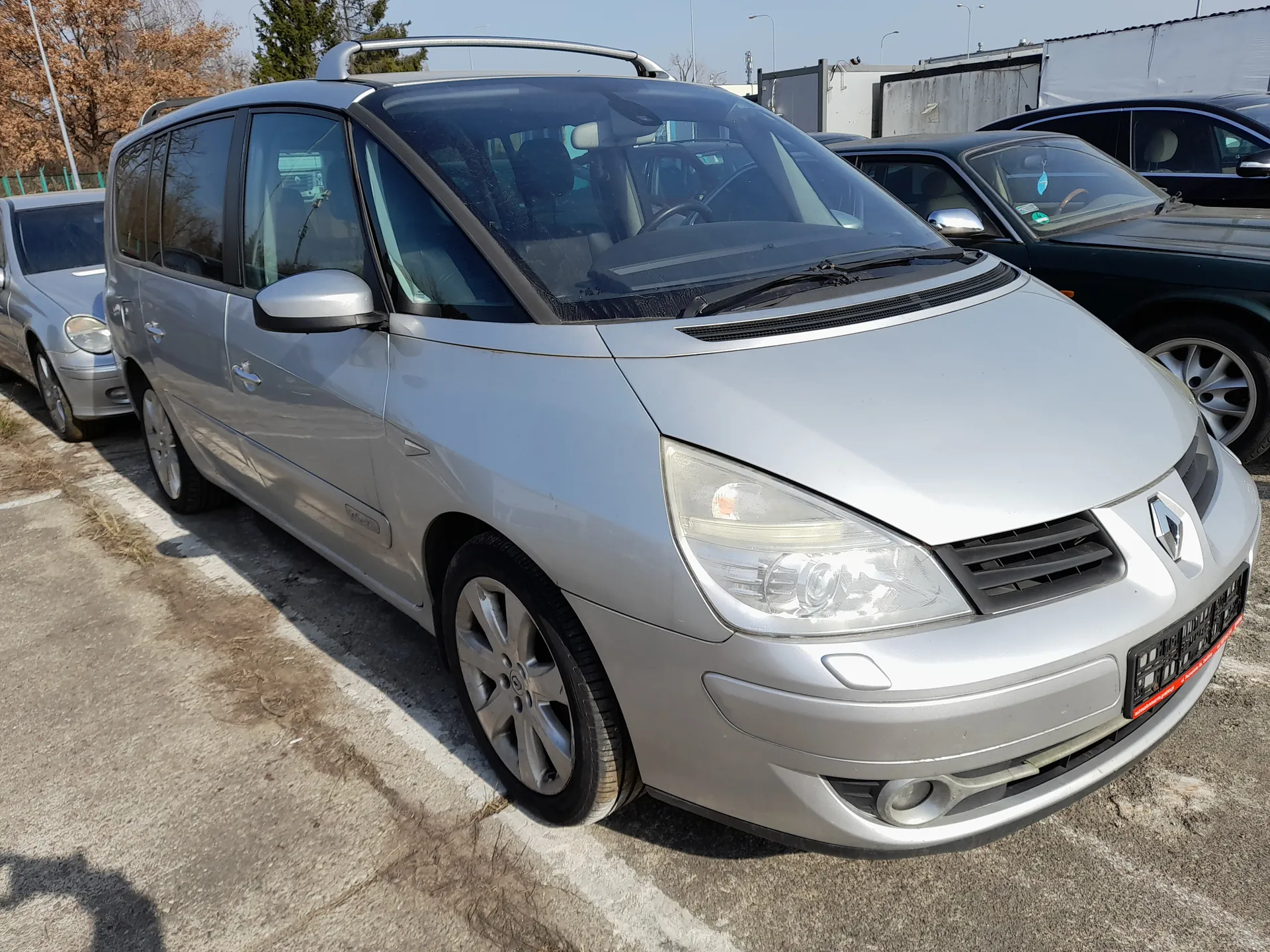 Srebrny minivan marki Renault z dachowym bagażnikiem zaparkowany na otwartym parkingu. W tle widoczne są inne pojazdy: srebrny sedan po lewej i ciemny samochód osobowy po prawej. Tło stanowią drzewa, ogrodzenie i budynki przemysłowe pod jasnym niebem. Na przednim zderzaku umieszczona jest tablica rejestracyjna z czerwonym napisem na dolnej krawędzi.
