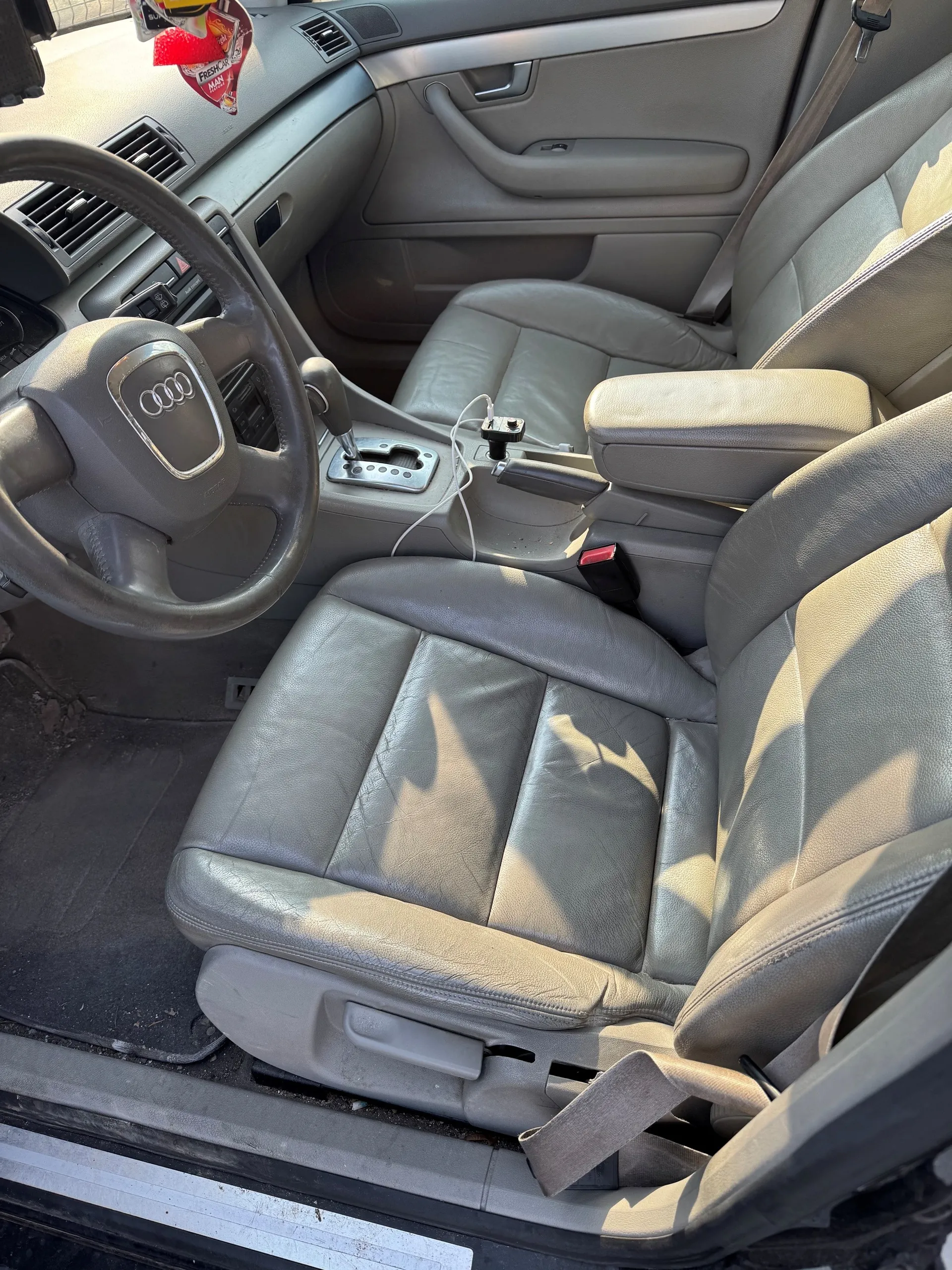 Wnętrze samochodu marki Audi z widoczną kierownicą z logo czterech pierścieni, szarymi skórzonymi fotelami, centralną konsolą z dźwignią zmiany biegów i białym kablem ładowania. Na desce rozdzielczej widoczne są wentylacyjne otwory i elementy sterujące. W tle drzwi pasażera z uchwytem i oknem. Na lusterku wstecznym wiszą czerwone zapachy. Podłoga pokryta czarną wykładziną, a drzwi kierowcy są otwarte.