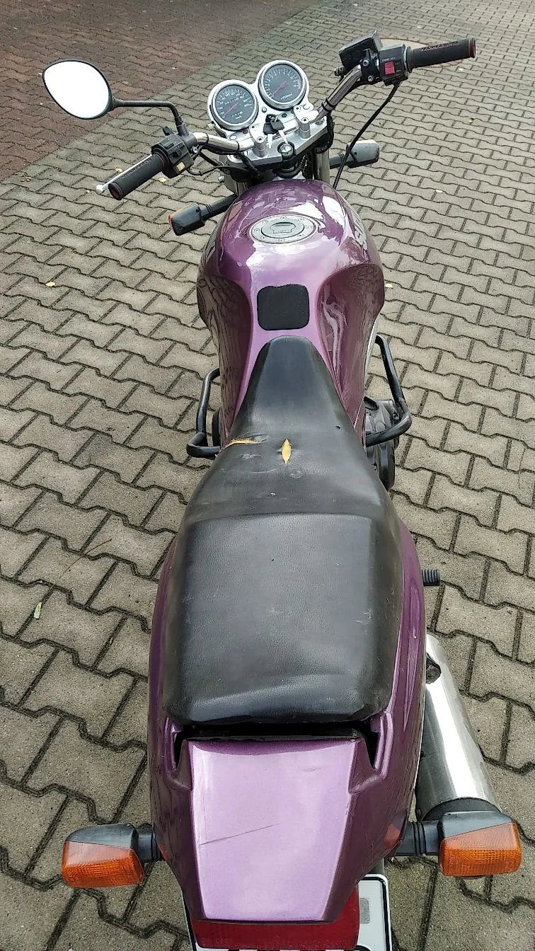Widok z góry na motocykl w kolorze fioletowym, zaparkowany na kostce brukowej. Widoczny jest czarny siedzenie z niewielkim uszkodzeniem materiału, fioletowy bak z wlewem paliwa, kierownica z lusterkiem po lewej stronie oraz dwa okrągłe zegary wskaźników. Z tyłu widoczne są pomarańczowe kierunkowskazy i fragment srebrnego wydechu.