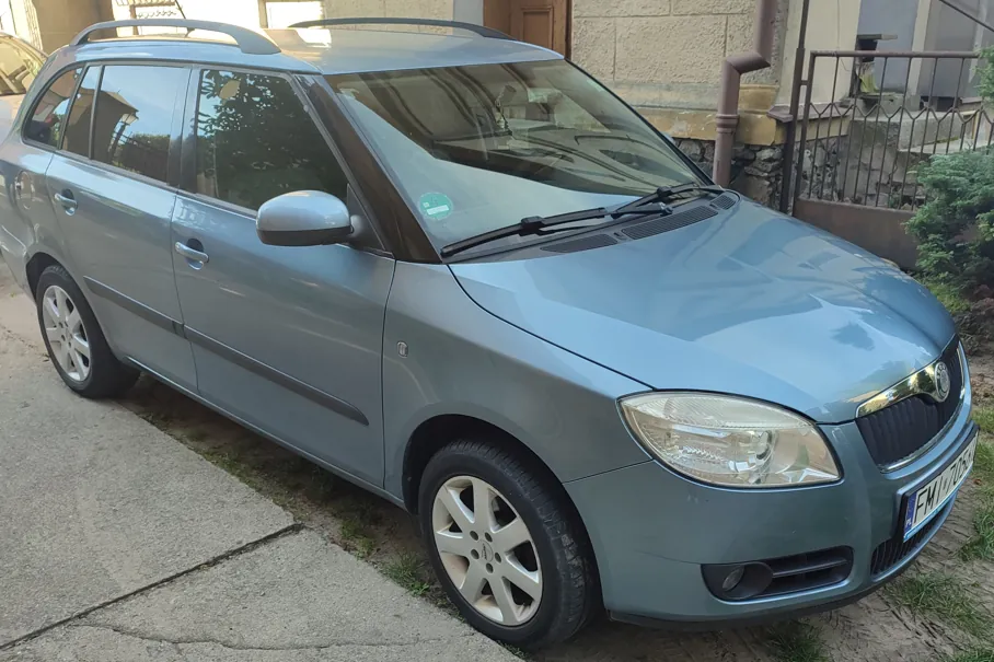 Jasnoniebieski samochód osobowy typu kombi zaparkowany na podjeździe przed budynkiem. Pojazd jest widoczny z przodu i z boku, posiada srebrne felgi aluminiowe oraz relingi dachowe. W tle widoczny jest fragment domu z jasną elewacją oraz metalowy płot.