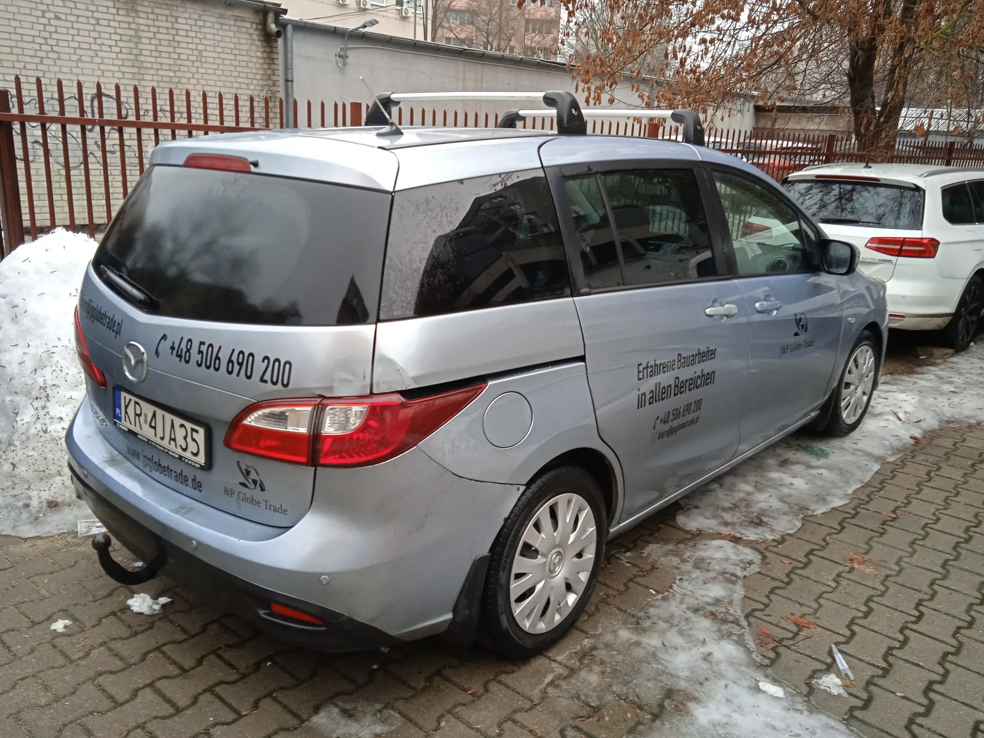 Srebrny samochód osobowy marki Mazda zaparkowany na brukowanej powierzchni z resztkami śniegu. Na tylnym szybie widoczny numer telefonu +48 506 690 200, tablica rejestracyjna KR 4JA35 oraz adres strony www.iglobetrade.de. Po bokach pojazdu napis w języku niemieckim: Erfahrene Bauarbeiter in allen Bereichen, numer telefonu +48 506 690 200 i nazwa firmy I&P Globe Trade. W tle biały samochód, ceglany mur, metalowe ogrodzenie oraz drzewa bez liści.