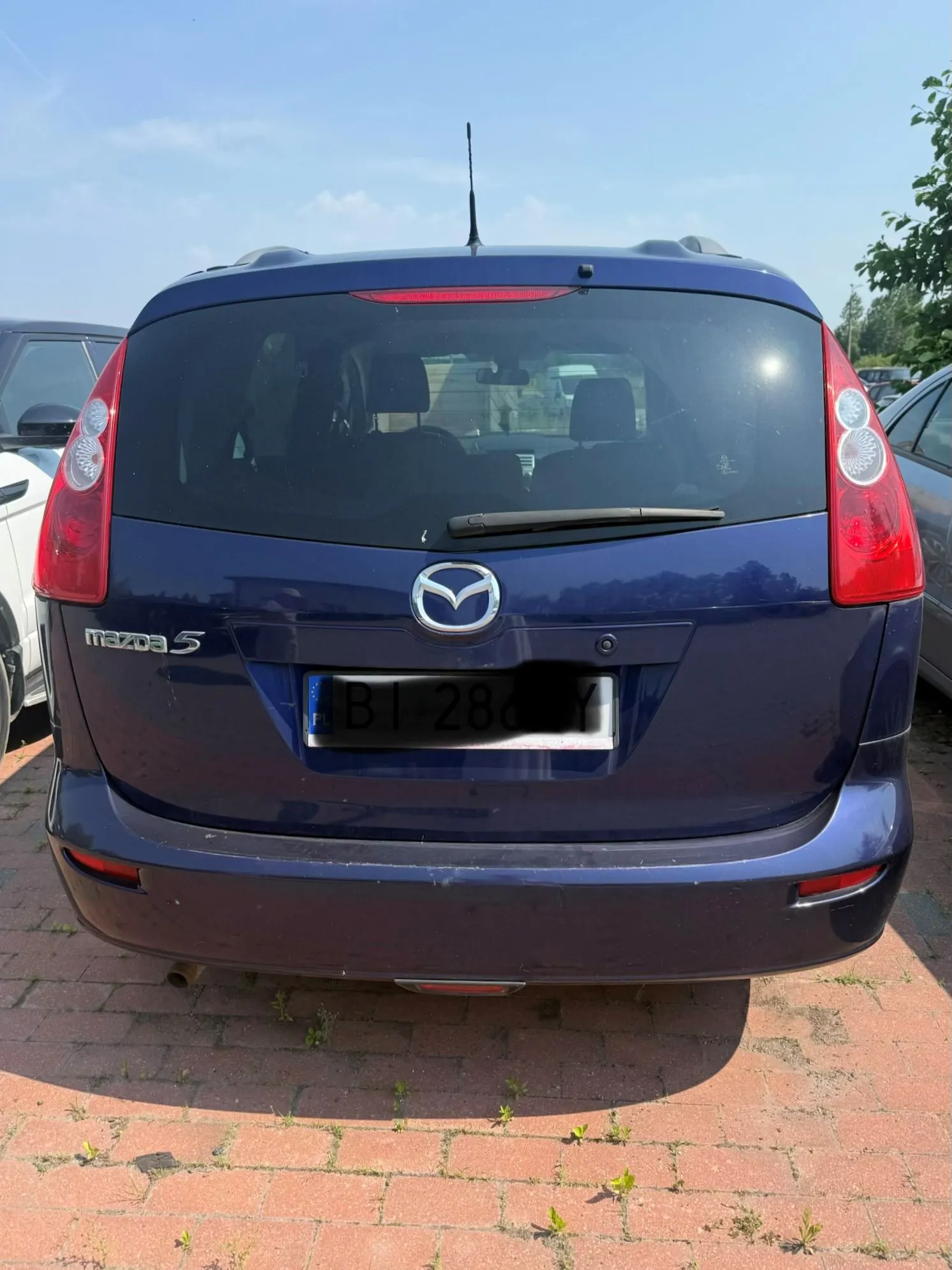 Tylny widok ciemnoniebieskiego samochodu marki Mazda 5 zaparkowanego na kostce brukowej. Na klapie bagażnika widoczne jest logo Mazdy, napis 'mazda 5' po lewej stronie oraz tablica rejestracyjna o treści 'B1 286 YY'. Samochód posiada tylną szybę z wycieraczką i antenę na dachu.
