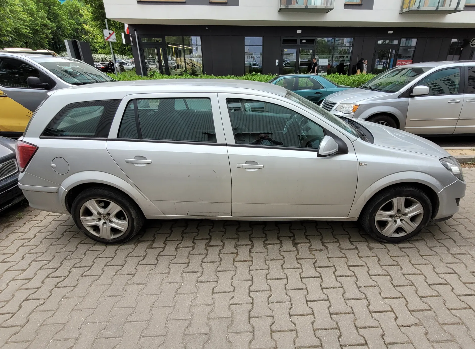 Srebrny samochód typu kombi zaparkowany na brukowanym placu. W tle widoczne są inne pojazdy: żółty samochód po lewej stronie, turkusowy i srebrny po prawej. Za nimi budynek z dużymi szybami i wejściem, przed którym stoją osoby. Po lewej stronie znak drogowy zakazujący wjazdu. Na pierwszym planie brukowane nawierzchnia parkingu.