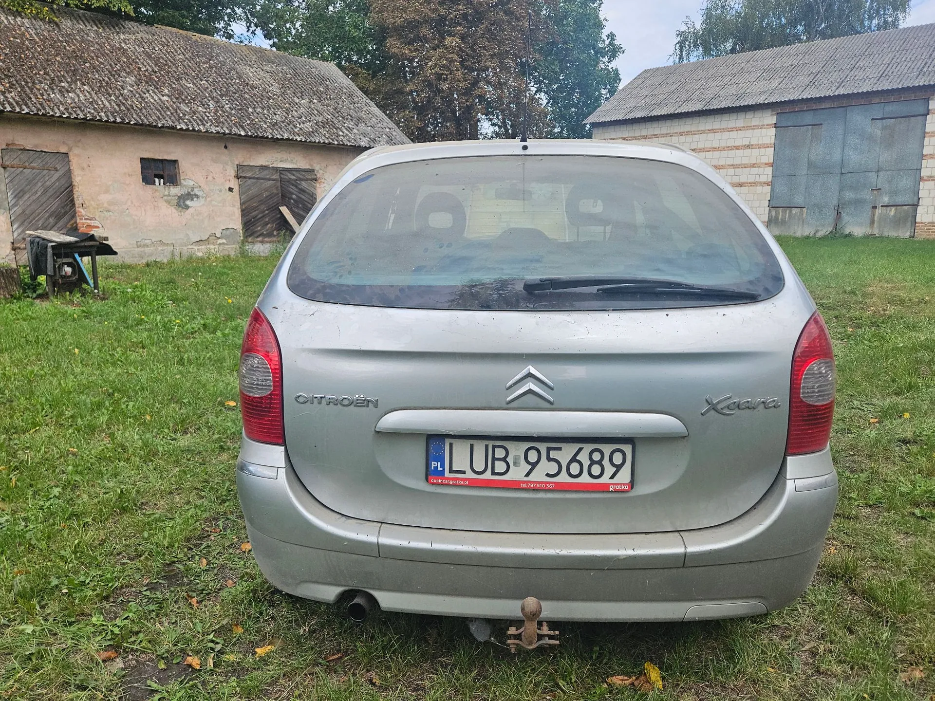 Tylny widok srebrnego samochodu marki Citroën Xsara zaparkowanego na trawiastym terenie. Na klapie bagażnika widoczne jest logo marki, napis CITROËN oraz model Xsara. Samochód posiada polską tablicę rejestracyjną o numerze LUB 95689. W tle znajdują się dwa stare budynki gospodarcze z dachami krytymi eternitem. Samochód jest wyposażony w hak holowniczy.