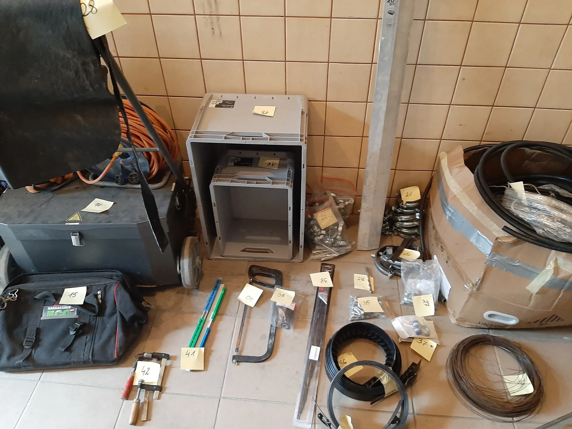 Zestaw narzędzi i przedmiotów ułożonych na podłodze z kafelkami. Po lewej stronie czarna walizka z żółtym znakiem ostrzegawczym i czarna torba z napisem 'HARDBASSER ECO' oraz etykietą '15'. W centrum szare plastikowe pojemniki, pomarańczowy przewód, piła ramiona, metalowe pręty, rolka drutu i różne małe elementy z żółtymi etykietami oznaczonymi numerami (np. 39, 41, 42). Po prawej stronie karton z czarnymi przewodami i opakowaniami. Tło stanowi beżowa ściana z kafelkami.