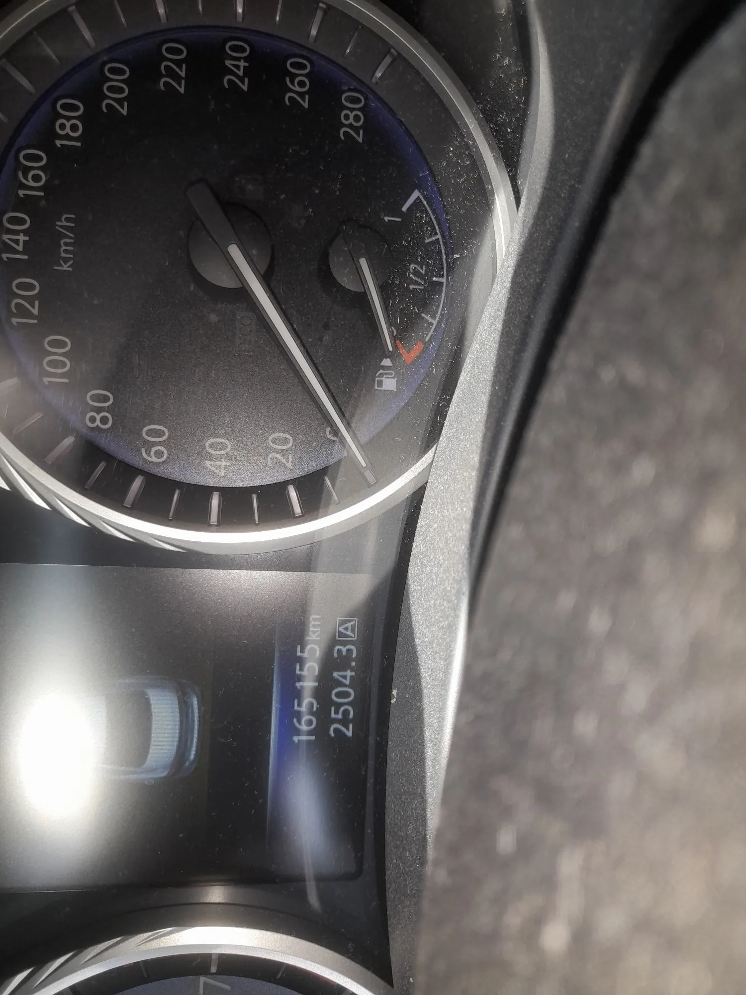 Zbliżenie na deskę rozdzielczą samochodu. Widoczny jest prędkościomierz z cyframi od 20 do 280 km/h oraz wskaźnik poziomu paliwa. Obok znajduje się cyfrowy wyświetlacz z tekstem: '165155 km' oraz '2504.3 A'. Obraz jest obrócony o 90 stopni w lewo.