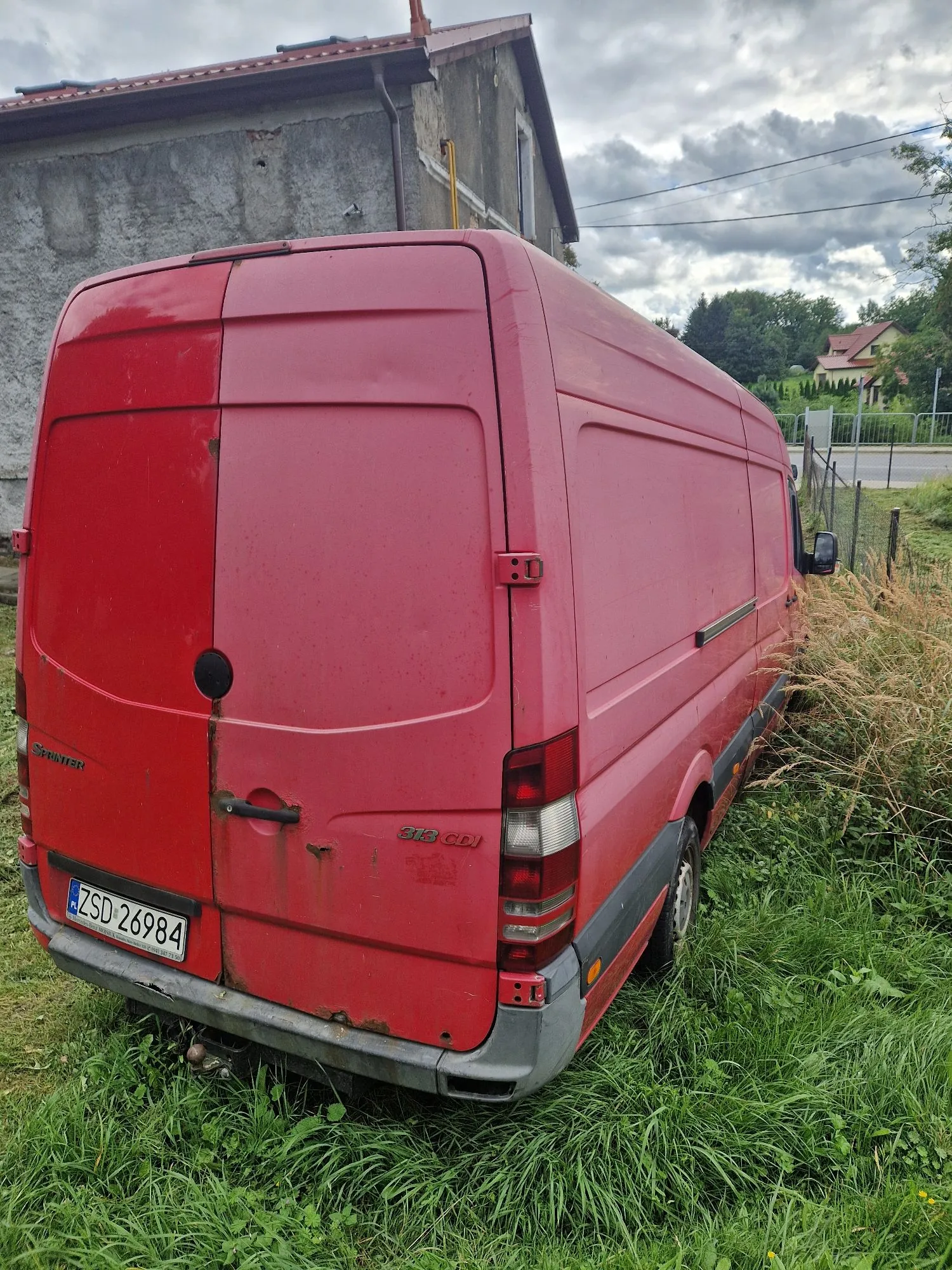 Tylny i boczny widok czerwonego vana marki Mercedes-Benz Sprinter, zaparkowanego w wysokiej trawie obok szarego budynku. Pojazd posiada widoczne ślady korozji na tylnych drzwiach oraz tablicę rejestracyjną o numerze ZSD 26984. Na tylnej klapie widnieją napisy 'SPRINTER' oraz '313 CDI'.