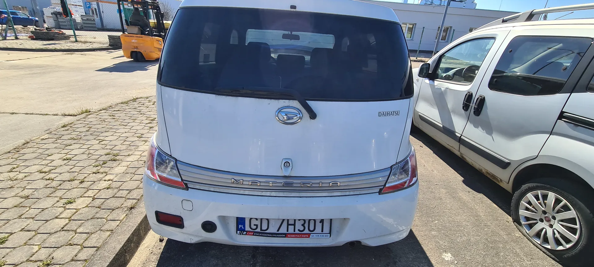 Biały samochód Daihatsu Materia z tablicą rejestracyjną GD 7H301 (PL) widziany od tyłu. Po prawej stronie biały samochód osobowy. W tle pomarańczowy wózek widłowy, budynki i brukowana powierzchnia. Na tylnym zderzaku widoczne napisy: DAIHATSU, MATERIA oraz tablica rejestracyjna z dodatkowym napisem STACJA KONTROLI POJAZDÓW.