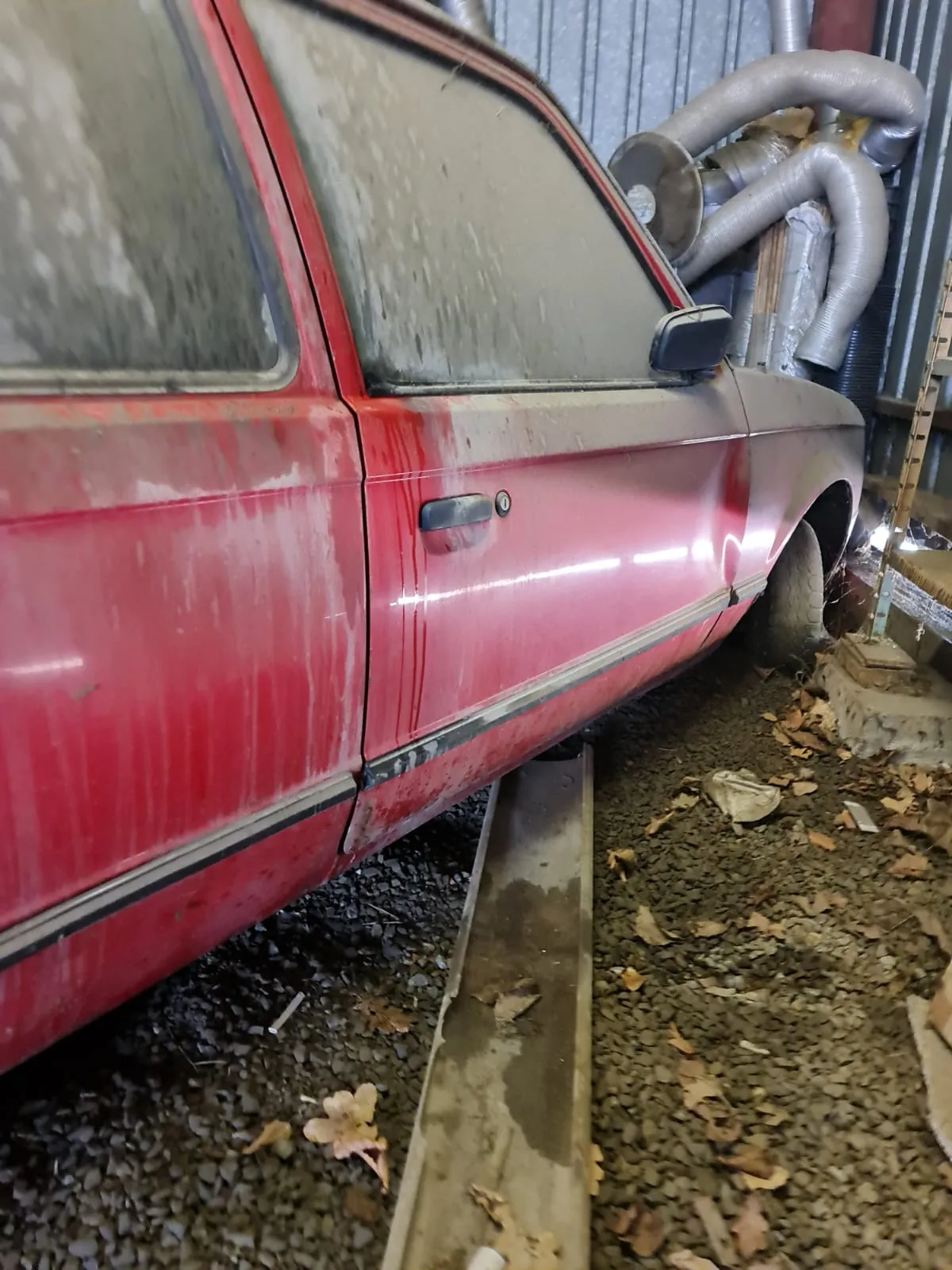 Czerwony samochód osobowy w stanie silnego zabrudzenia, z widocznymi śladami kurzu i brudu na karoserii oraz szybach. Samochód stoi na podłożu z drobnej kruszywy, wśród rozrzuconych liści. W tle metalowe rury i elementy konstrukcyjne, prawdopodobnie wewnątrz garażu lub hali. Widoczne są drzwi pojazdu z uchwytem oraz lusterko boczne.