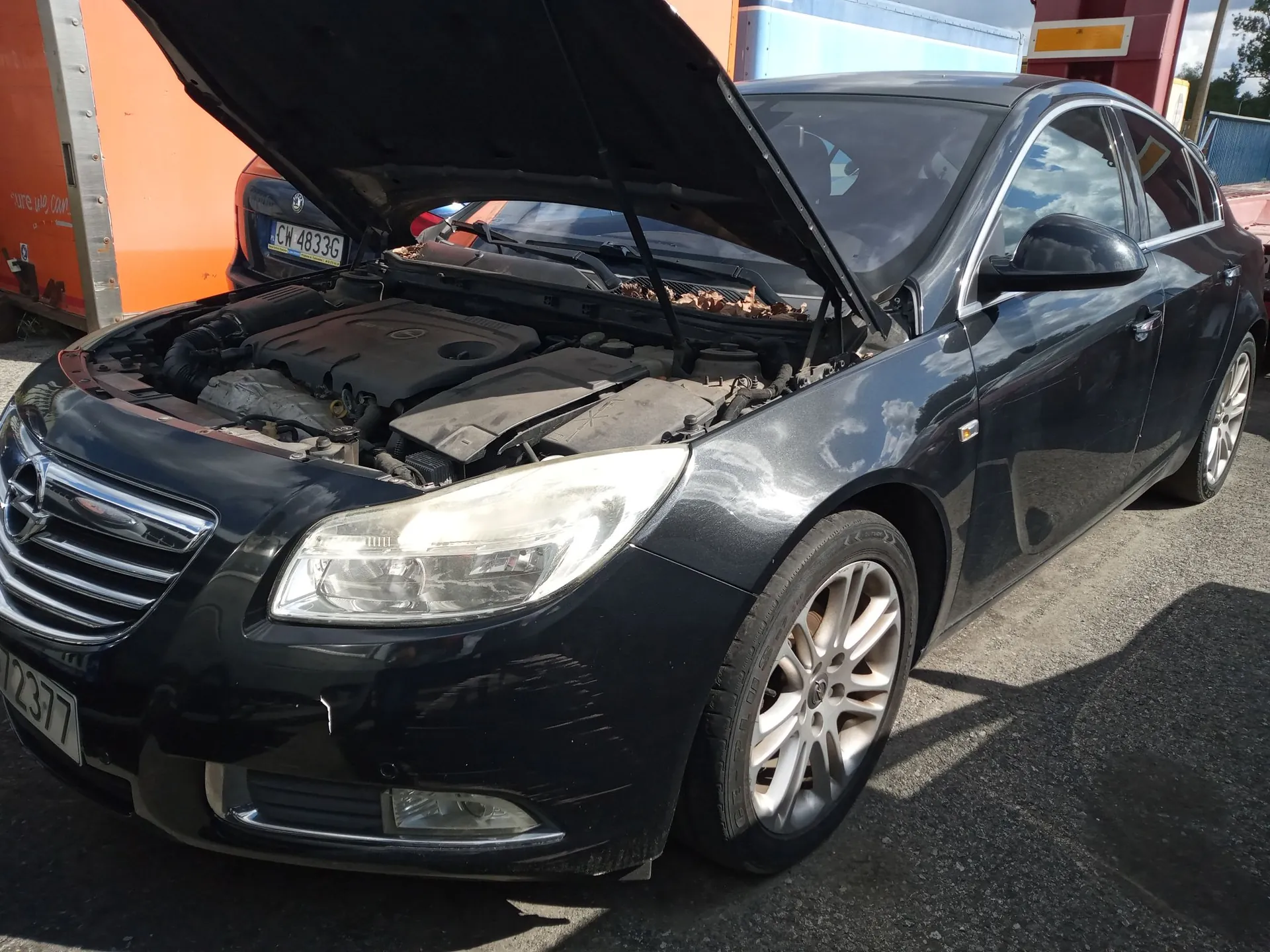 Czarny samochód marki Opel z otwartą maską silnika, zaparkowany na zewnątrz. Widoczny jest przód pojazdu z reflektorem, grillem i kołem. W tle znajduje się pomarańczowy kontener oraz inny samochód z tablicą rejestracyjną CW 4833G.