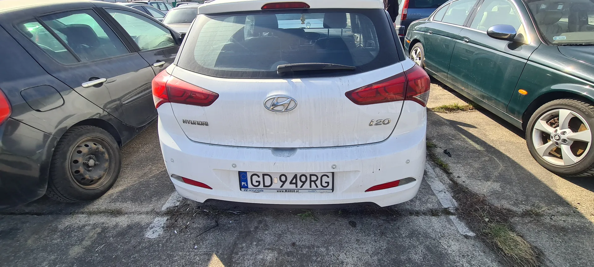 Biały samochód Hyundai i20 z tyłu, z widocznym logo marki, napisem HYUNDAI po lewej stronie bagażnika i oznaczeniem i20 po prawej. Tablica rejestracyjna PL GD 949RG z dodatkowym napisem www.MARGO.pl na dole. Po lewej stronie ciemnoszary samochód z brudnym kołem, po prawej zielony samochód. Tło stanowi parking z betonowym nawierzchniem i fragmentami trawy.