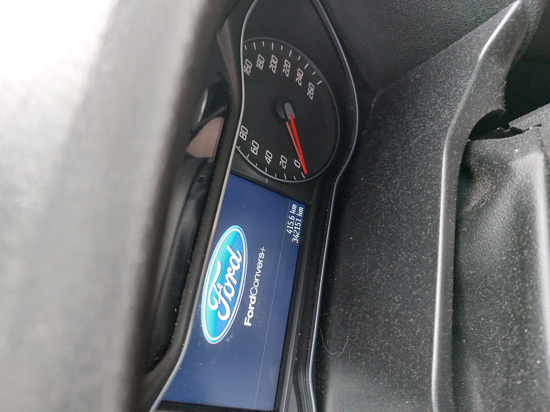 Zbliżenie na deskę rozdzielczą samochodu marki Ford. Widoczny jest analogowy prędkościomierz z podziałką do 260 km/h oraz niebieski ekran cyfrowy z logo Ford i napisem 'FordConvers+'. Na ekranie widoczne są liczby: 615,6 km oraz 342151 km.