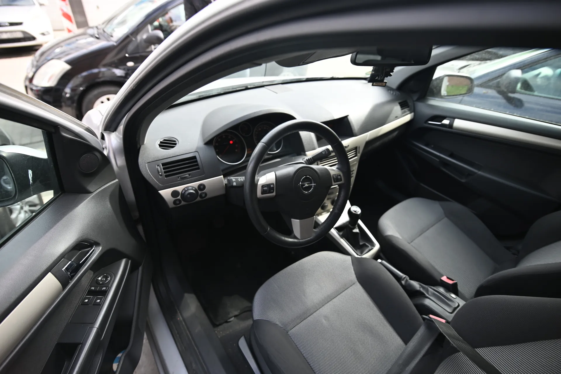 Wnętrze samochodu marki Opel z otwartą lewą przodem drzwi. Widoczna kierownica z logo Opel, deska rozdzielcza z licznikami, szare siedzenia z czarnymi akcentami, dźwignia zmiany biegów oraz drzwi z przyciskami sterowania szybami. Na zewnątrz przez otwarte drzwi i przednią szybę widoczne są inne zaparkowane samochody.