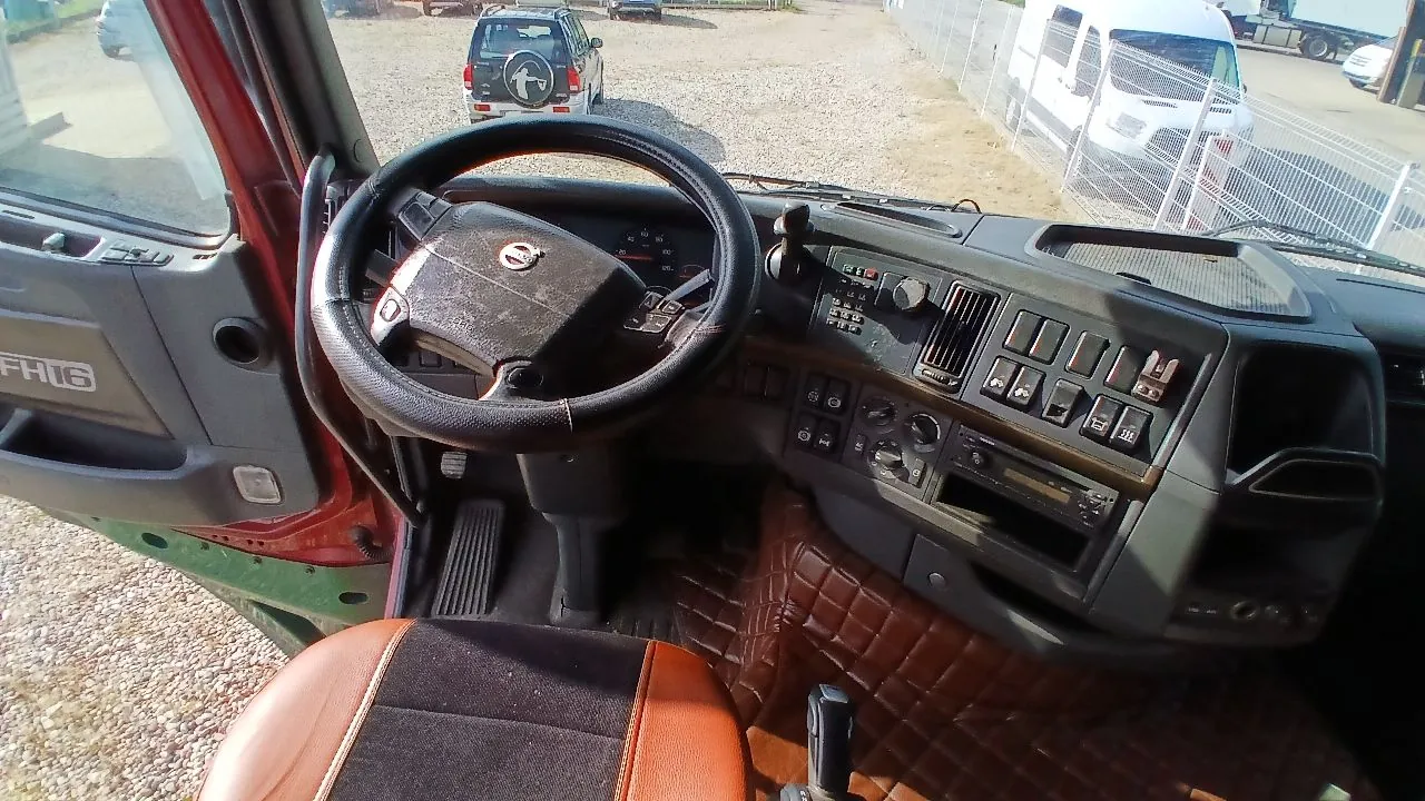 Widok z wnętrza kabiny ciężarówki, z perspektywy kierowcy. Widoczna jest czarna kierownica z logo Volvo, deska rozdzielcza z licznikiem, licznymi przełącznikami i przyciskami oraz lewy panel drzwi z napisem 'FH16'. W dolnej części zdjęcia widoczny jest fragment brązowego fotela oraz brązowa, pikowana wykładzina na podłodze. Przez przednią szybę widać parking z zaparkowanym białym samochodem terenowym i białym furgonetem.