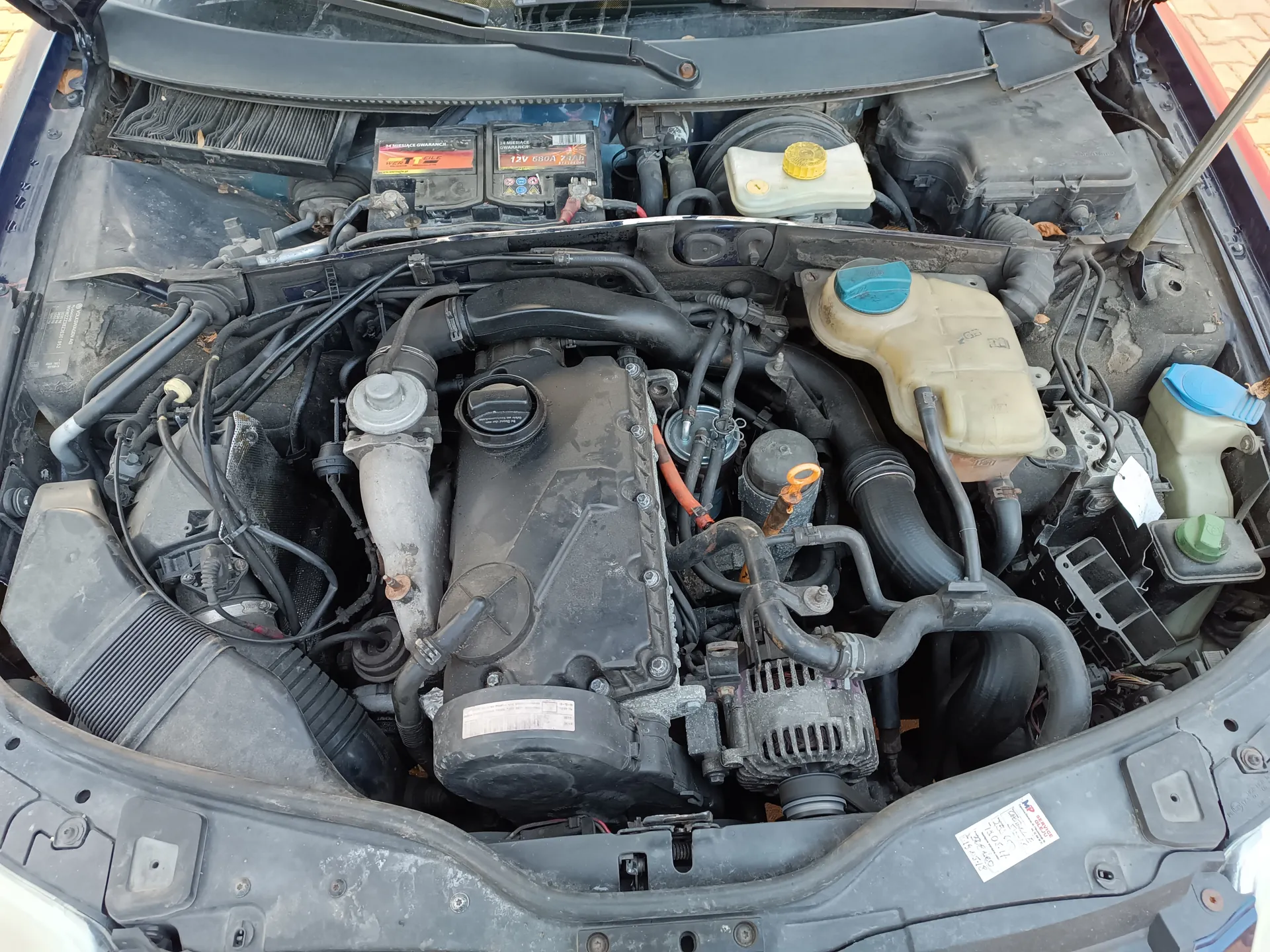 Widok z góry na komorę silnika samochodu z otwartą maską. Centralnie umieszczony silnik, liczne przewody i węże. Widoczne zbiorniki z kolorowymi korkami: żółty, niebieski i zielony. Bateria z dwiema etykietami zawierającymi napisy '12V 50Ah 450A'. Zanieczyszczony filtr powietrza po lewej stronie. Na prawej stronie komory silnika naklejka z nieczytelnym tekstem. Całość pokryta kurzem i śladami zużycia.