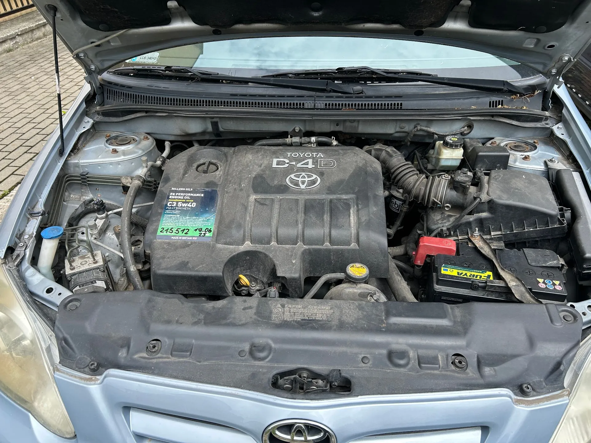 Widok z góry na komorę silnika w jasnoniebieskim samochodzie marki Toyota. Na plastikowej osłonie silnika widnieje napis 'TOYOTA D-4D'. Na osłonie przyklejona jest etykieta z informacją o oleju 'MILLERS TRILLS C3 5w40' oraz odręcznym zapisem '215512 18.06.23'. Po prawej stronie znajduje się czarny akumulator marki FURYA. Maski samochodu jest otwarta i podparta.