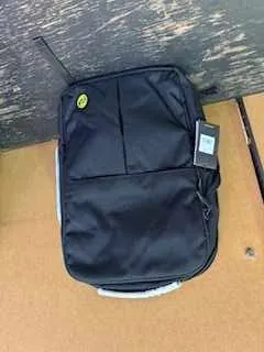 Czarny plecak z przednią kieszenią i żółtym logo w kształcie uśmiechu. Plecak leży na brązowej powierzchni kartonu oraz ciemnym, zużytym drewnianym podłożu. Na plecaku widoczna jest biała etykietka.