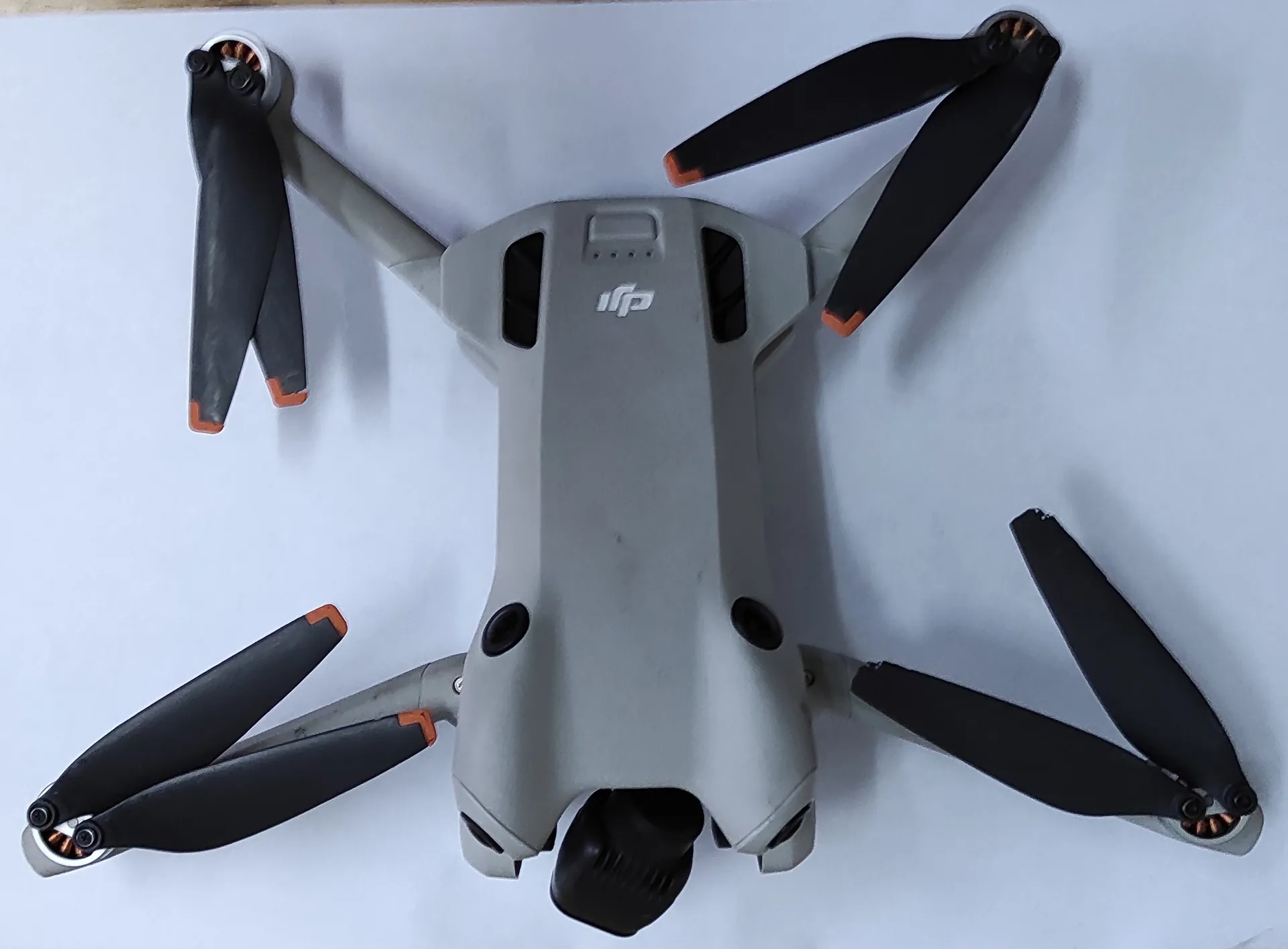 Szary dron marki DJI z czterema czarnymi śmigłami z pomarańczowymi końcówkami, leżący na białym tle. Na górnej obudowie widoczne jest białe logo DJI.