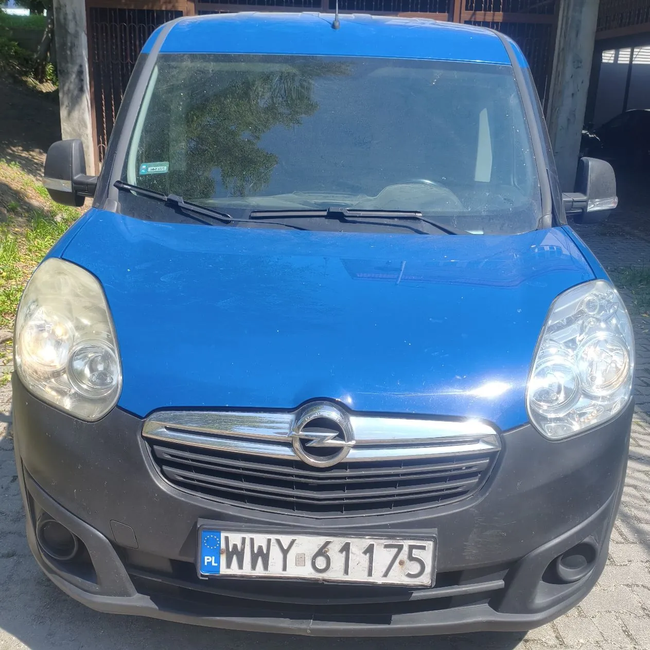 Niebieski samochód marki Opel widziany od przodu. Pojazd ma czarny zderzak i polską tablicę rejestracyjną o numerze WWY 61175. Lewy reflektor jest zmatowiały.