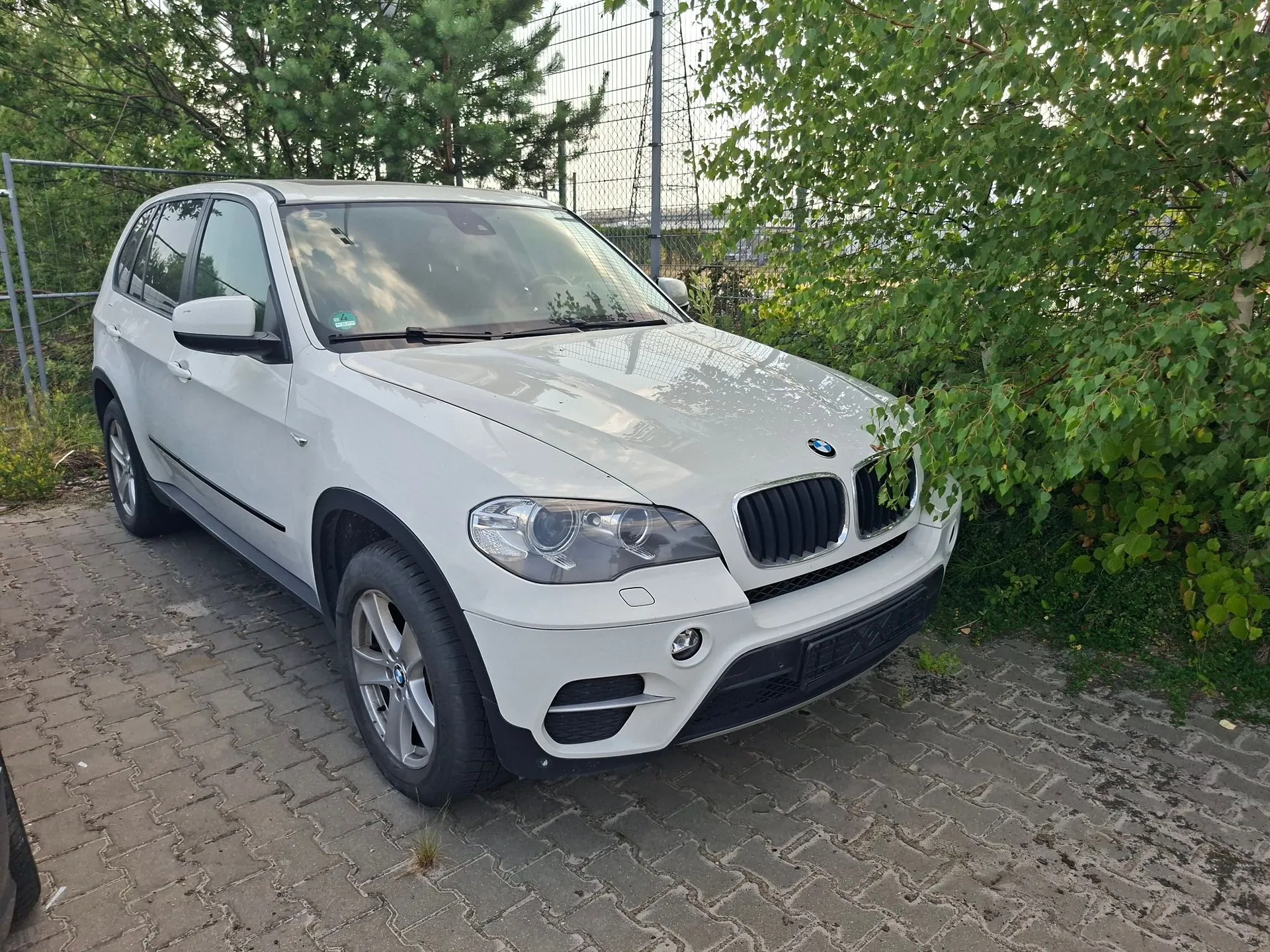 Biały samochód marki BMW typu SUV zaparkowany na kostce brukowej. Pojazd jest ustawiony pod kątem, widoczny z przodu i z lewej strony. W tle znajduje się metalowy płot oraz zielone drzewa i krzewy.
