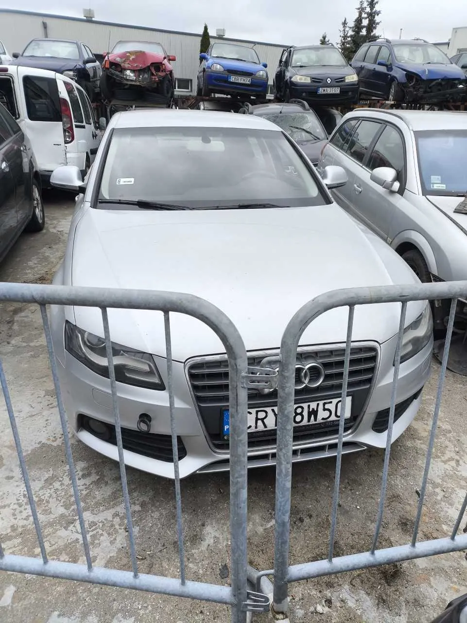 Srebrny samochód marki Audi z przodu, z widoczną tablicą rejestracyjną CRY 8W50. Przed pojazdem metalowa bariera. W tle inne samochody, w tym uszkodzone (czerwony z zniszczonym przodem) oraz zaparkowane na platformie. Po lewej stronie biały van, po prawej srebrny samochód. W tle budynek przemysłowy i drzewa. Niebo zachmurzone.