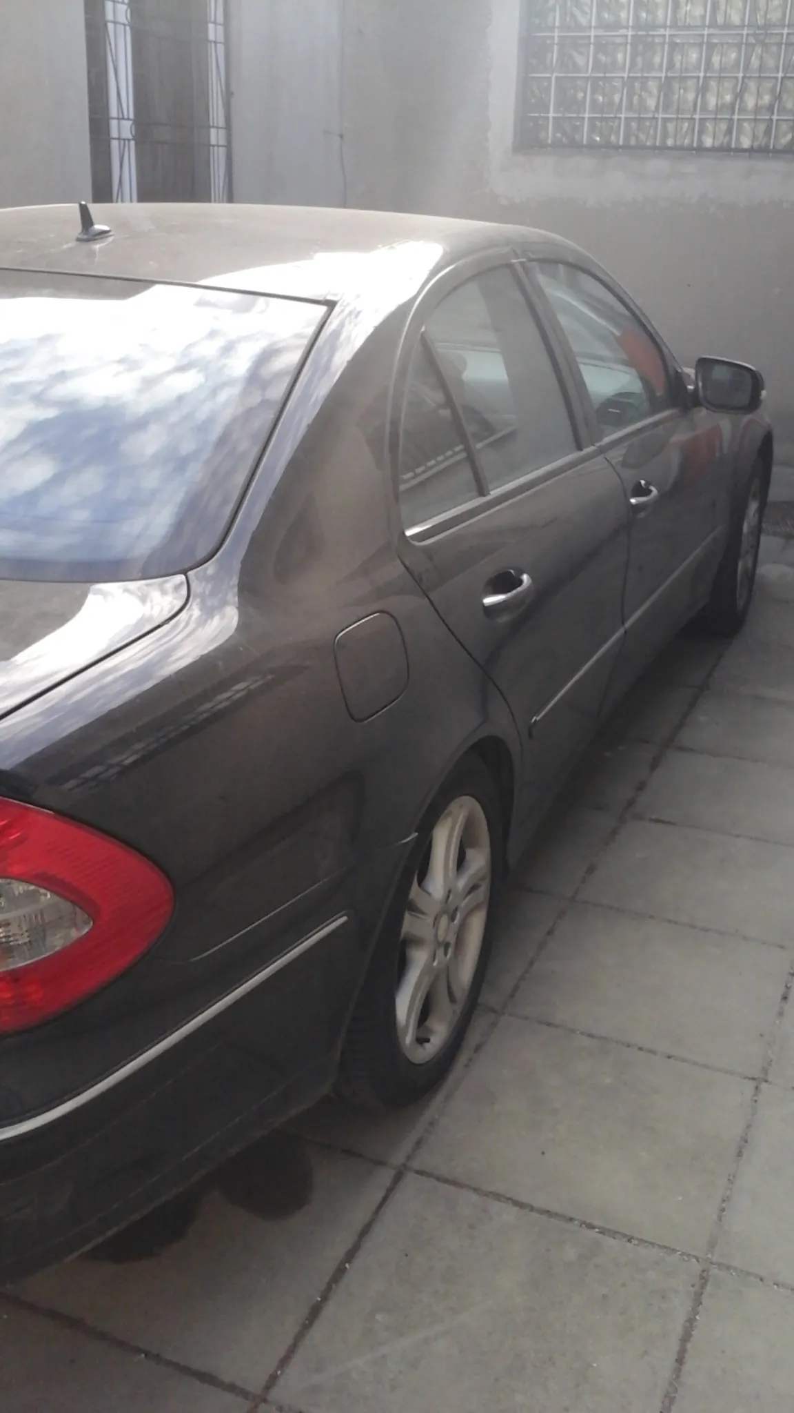 Ciemnoszary samochód osobowy zaparkowany przy betonowej ścianie z dużymi płytkami. Widoczna tylna część pojazdu z czerwonym światłem tylnym, kołem oraz lusterkiem bocznym. Po lewej stronie obrazu znajduje się okno z metalową kratą i szklanymi blokami. Na ścianie widoczny cień osoby.