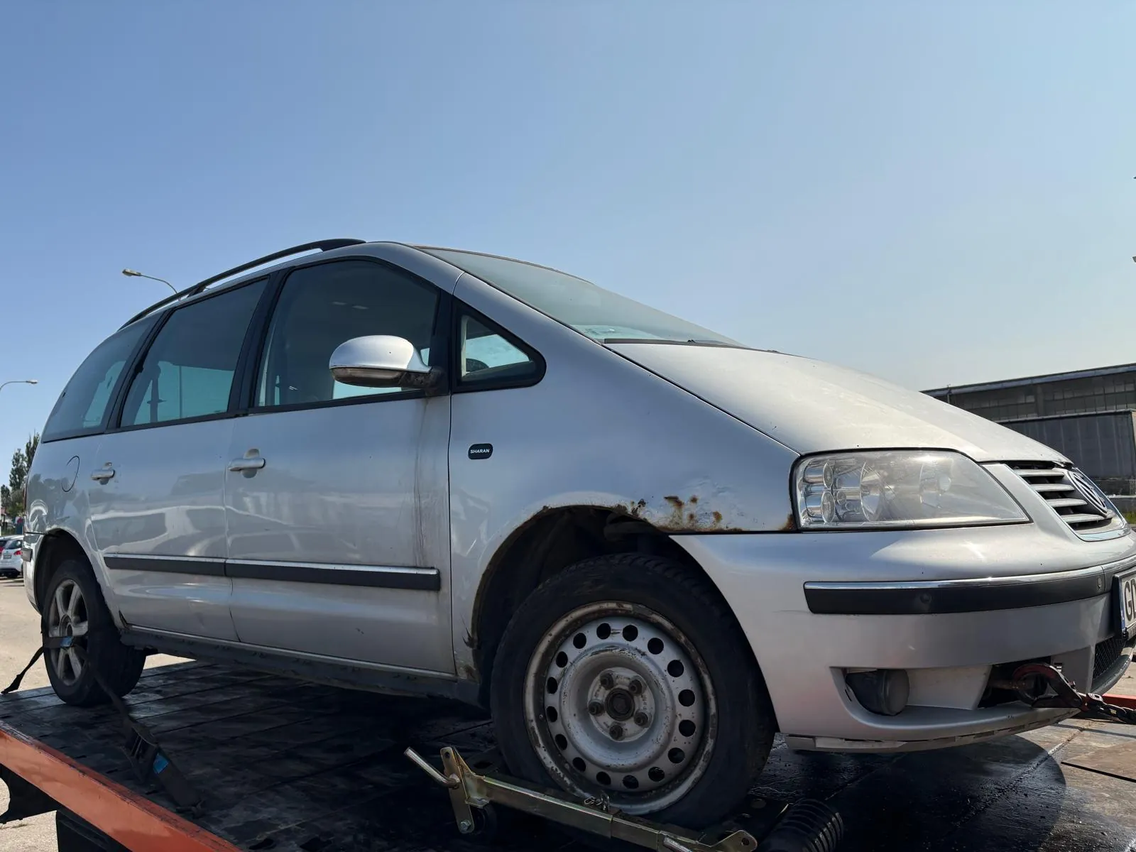 Srebrny minivan Volkswagen Sharan na platformie holownika. Samochód ma widoczne ślady rdzy na przednim lewym błotniku i kołach. W tle błękitne niebo oraz przemysłowa budowla. Minivan wyposażony w dachowy bagażnik i czarne pasy dekoracyjne wzdłuż boku. Koła z stalowymi felgami z otworami. Platforma holownika pomarańczowo-czarna, samochód przytwierdzony pasami.