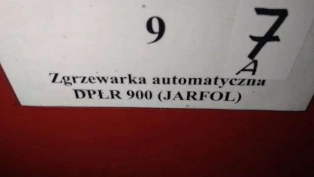 Biała tabliczka z czarnym tekstem. Na górze znajduje się cyfra 9, a po prawej stronie mniejsza naklejka z cyfrą 7 i literą A poniżej. Poniżej cyfry 9 widnieje napis: 'Zgrzewarka automatyczna DPŁR 900 (JARFOL)'. Tabliczka znajduje się na czerwonym tle.