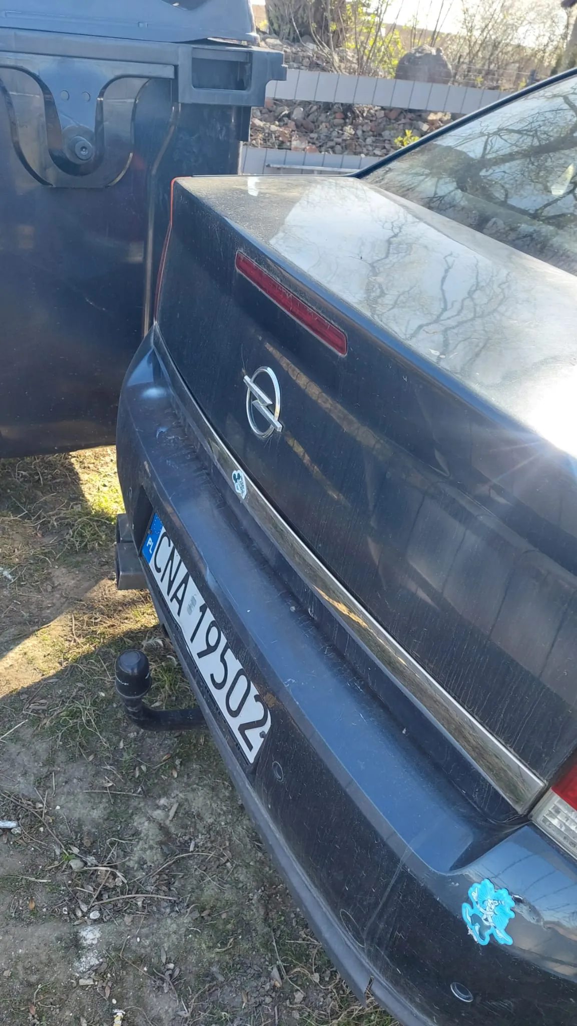 Tył czarnego samochodu marki Opel z widocznym logo na bagażniku. Tablica rejestracyjna z białym tłem i czarnym napisem CNA 19502 oraz niebieskim polem z literami PL po lewej stronie. Na bagażniku znajdują się dwa naklejki: jedna biała w kształcie postaci i niebieska z motywem zwierzęcym w prawym dolnym rogu. Samochód stoi na trawiastym i ziemnym terenie obok dużego czarnego pojemnika na śmieci. W tle widoczny jest kamienny mur, suche drzewa i fragment ogrodzenia.